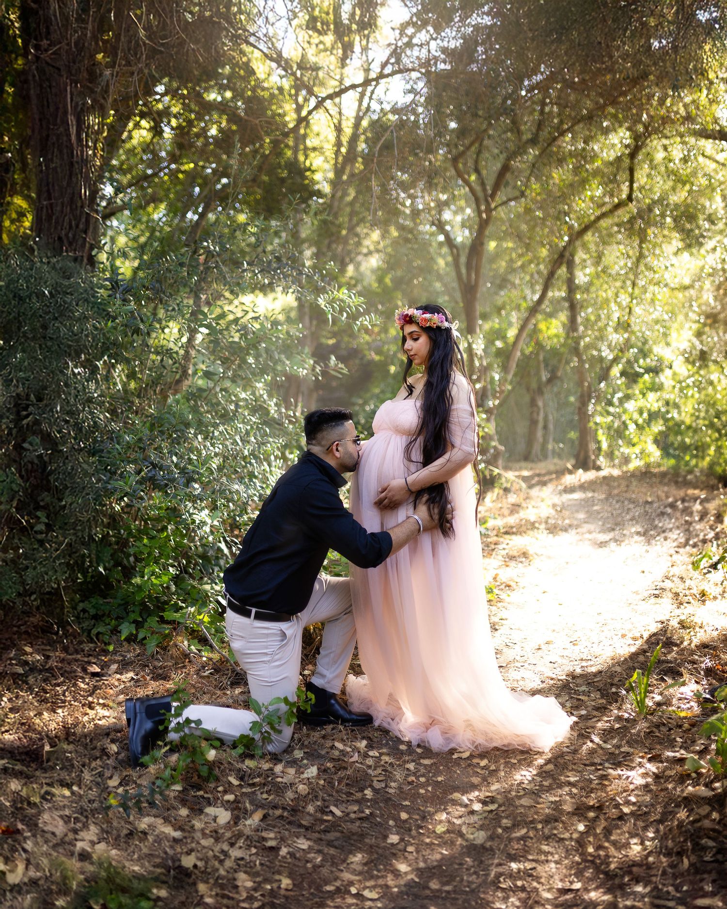A romantic couple shares tender moments in a sunlit forest clearing while wearing formal attire.