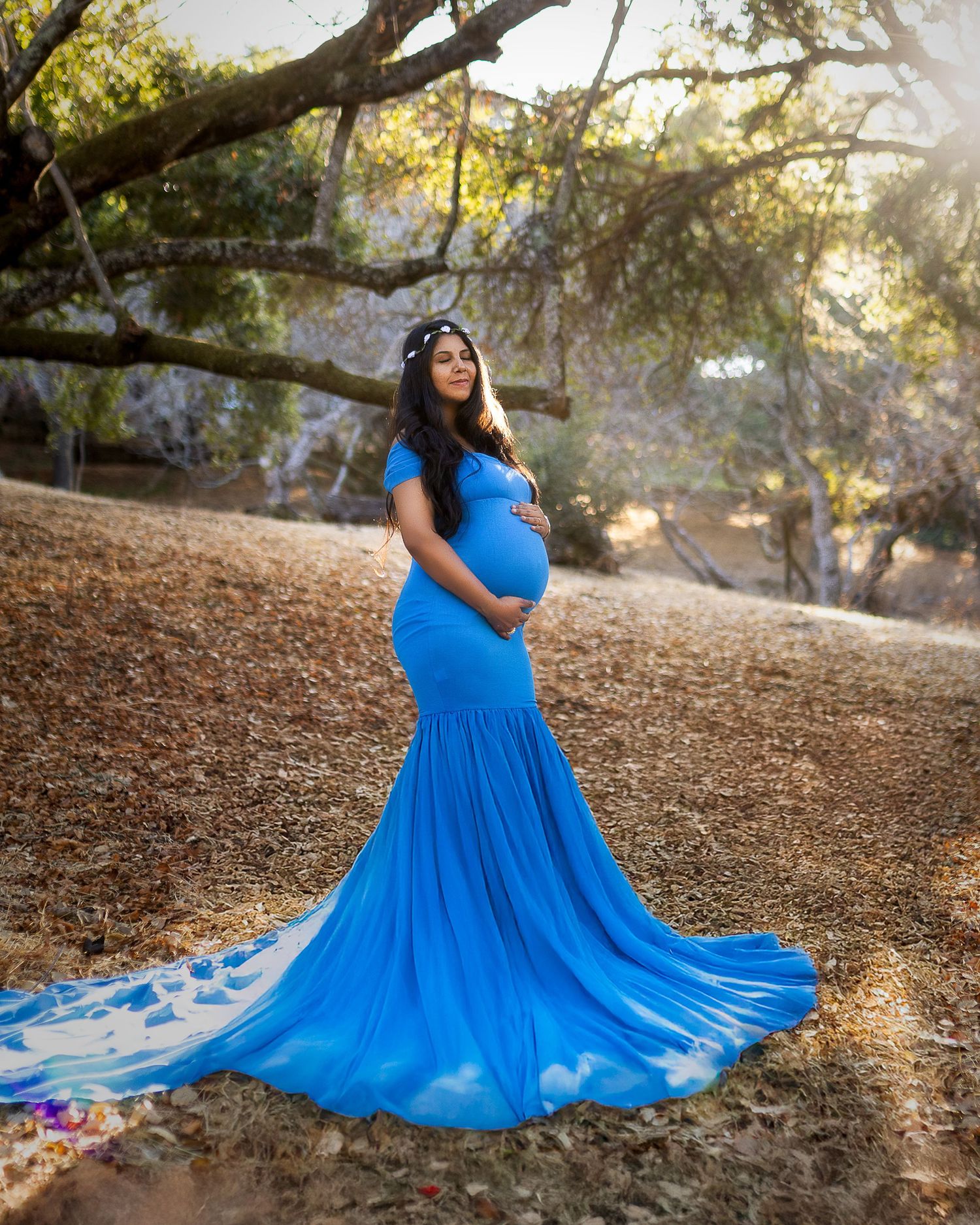 A bright blue flowy dress spreads out dramatically against fallen leaves under a large tree.