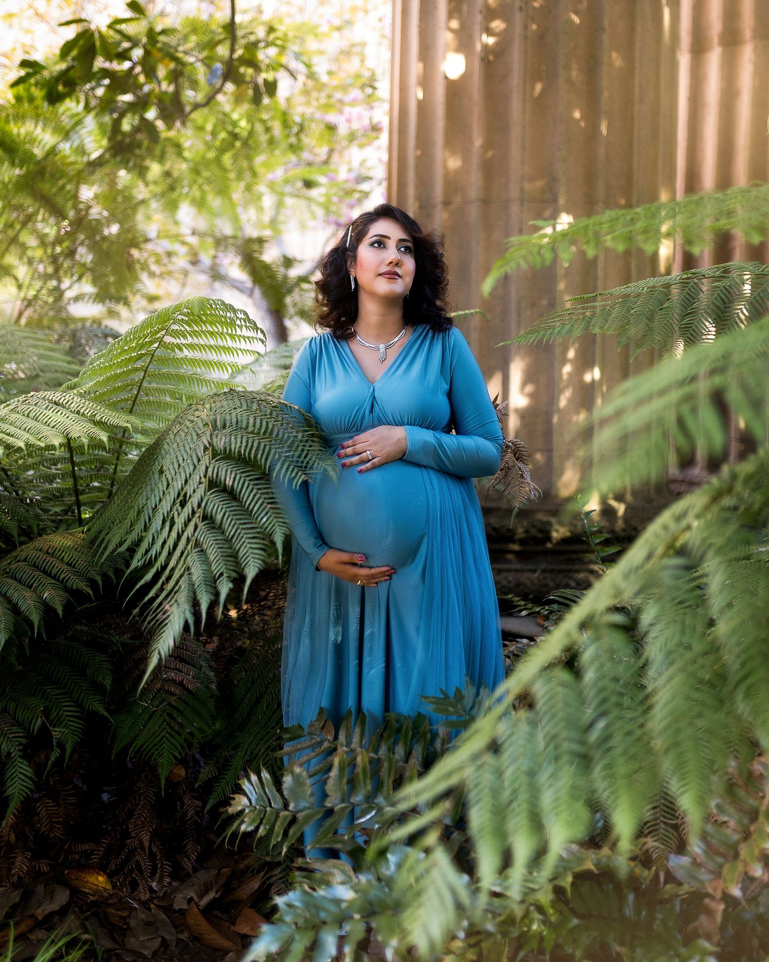 A figure in a flowing blue dress poses elegantly among lush green fern leaves in a sunlit outdoor setting.