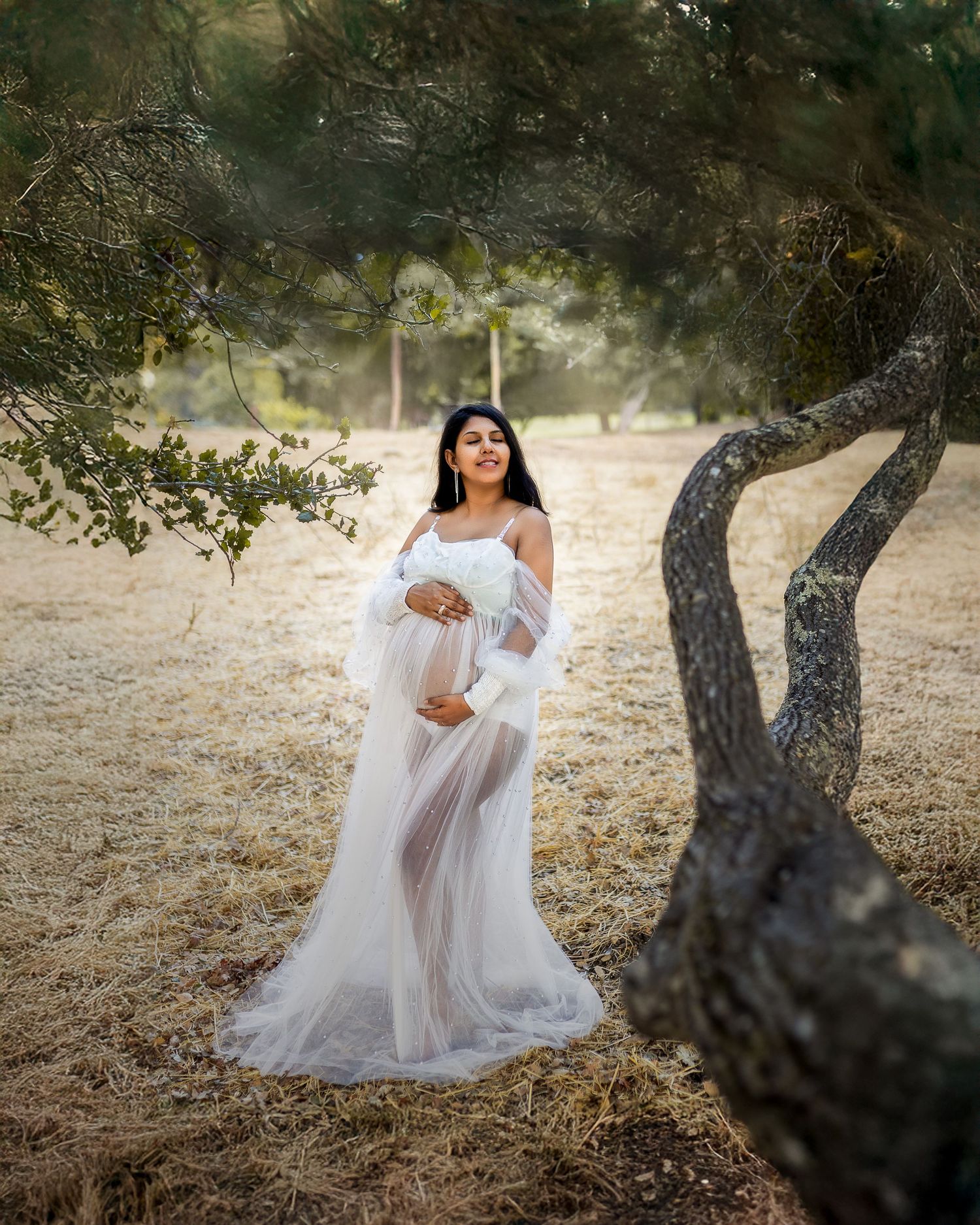 A dreamy outdoor maternity photoshoot with a flowing white dress against a backdrop of rustic trees and natural light.