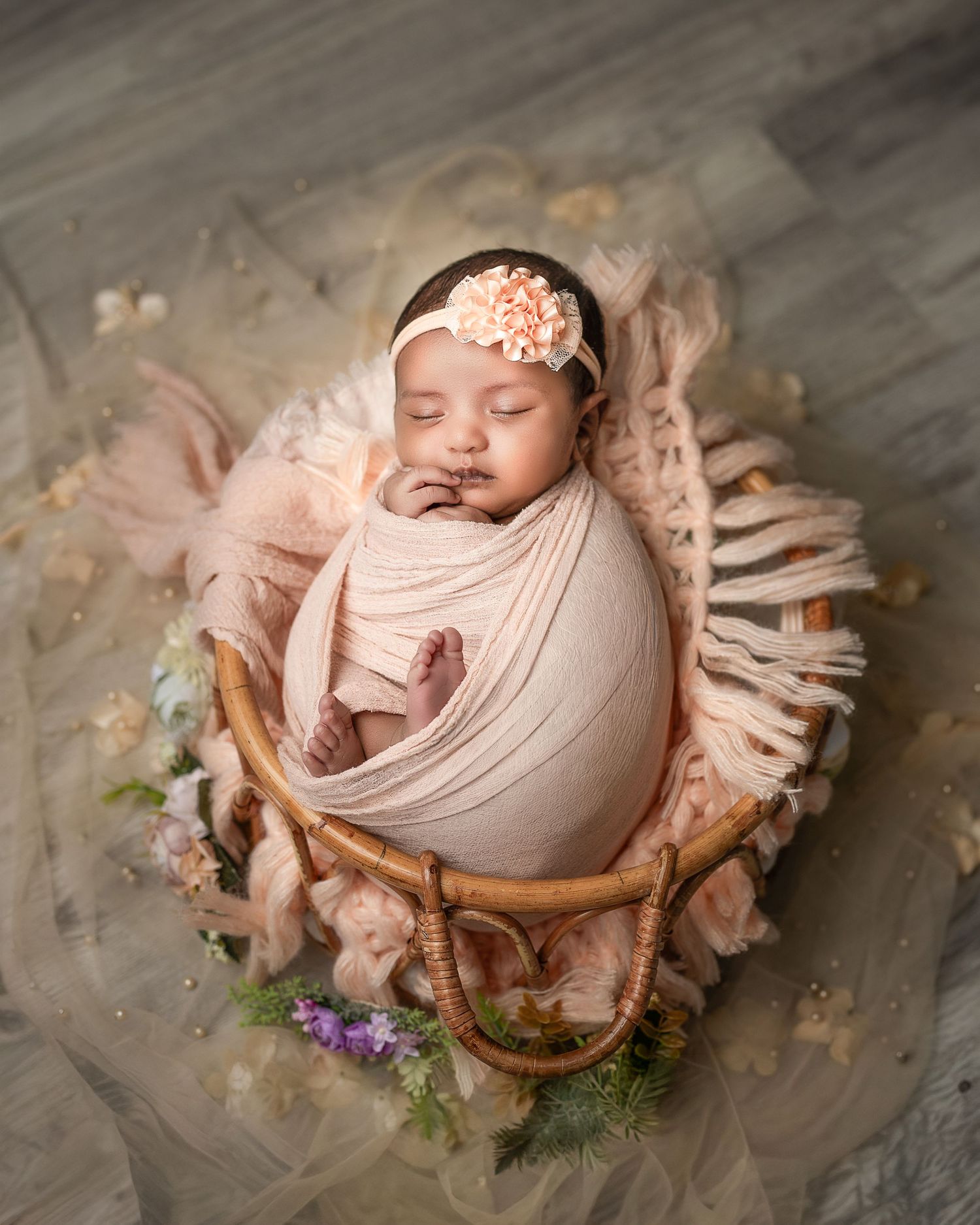 Newborn sleeping in a rustic wooden bowl surrounded by soft pink fabric and delicate flowers.