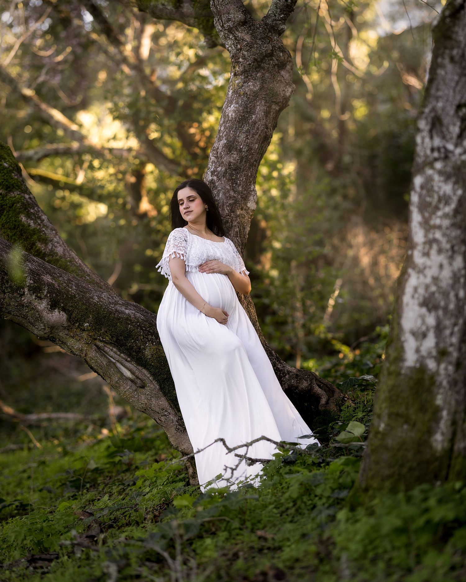 Person in flowing white dress sits gracefully on mossy tree branches in enchanted forest setting during golden hour.