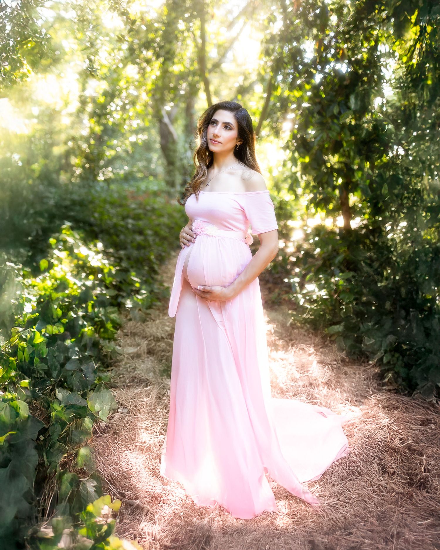 A dreamy outdoor maternity photoshoot featuring a pink flowy dress against sunlit foliage in a garden setting.