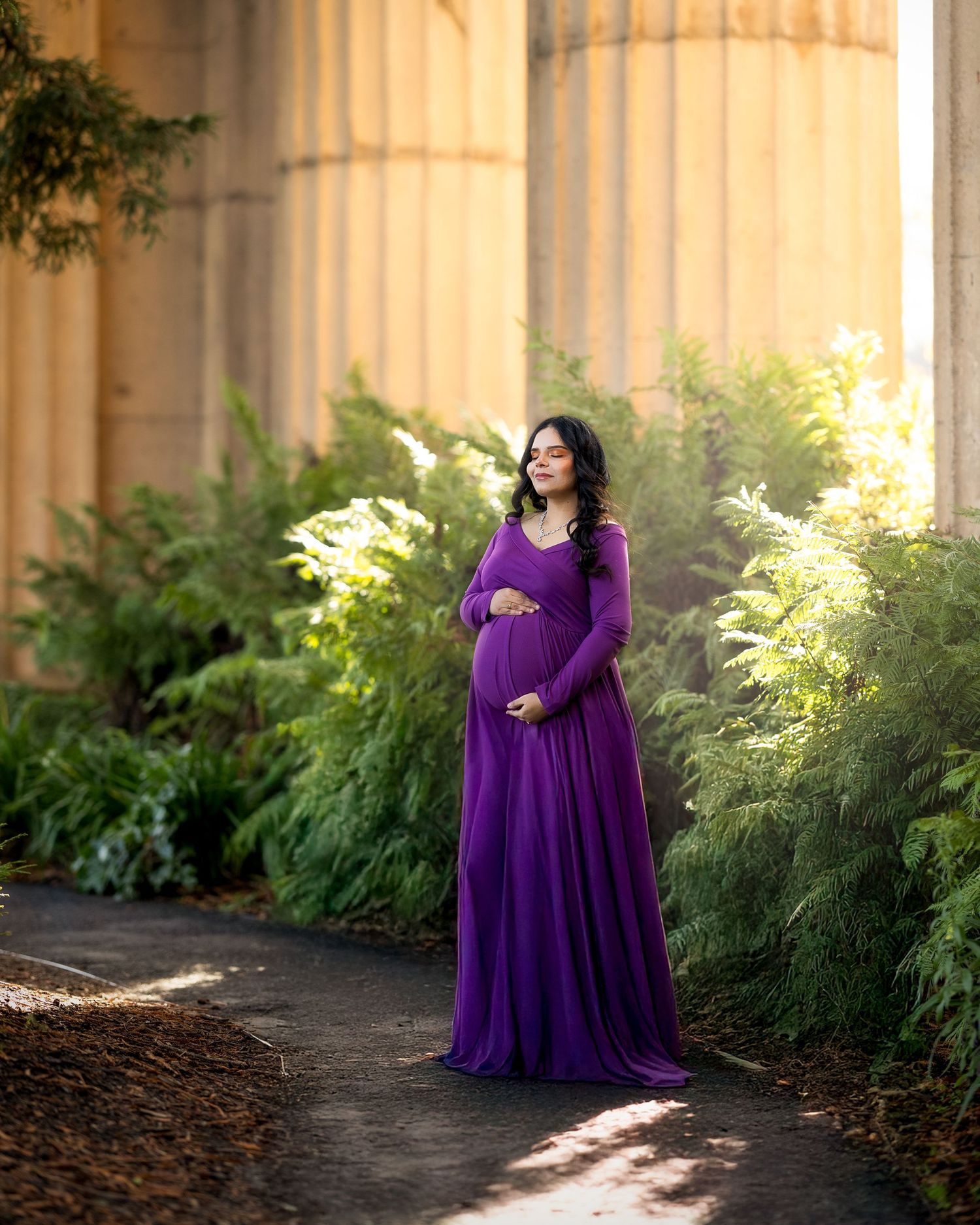 A pregnant woman in a flowing purple gown stands gracefully in front of stone columns with sunlight streaming through.