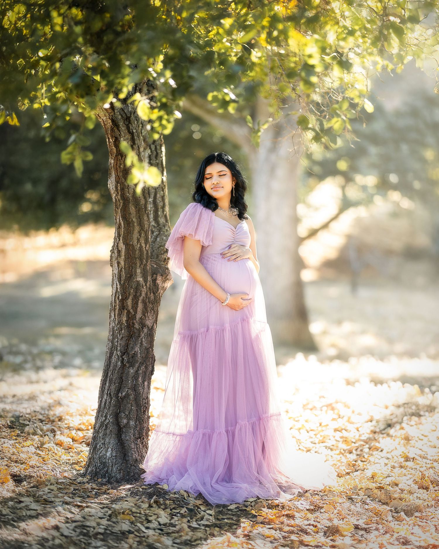 Ethereal maternity portraits in a sun-dappled forest setting with flowing lavender gowns and golden autumn light.
