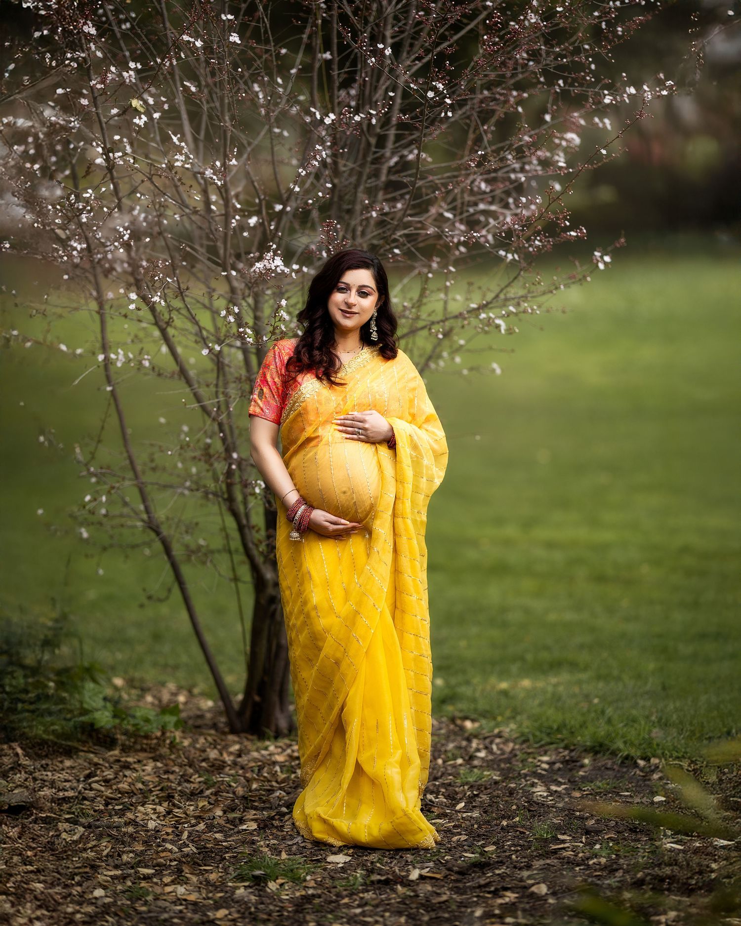 A bright yellow saree-clad figure stands by a blooming spring tree with delicate white blossoms in a green garden setting.