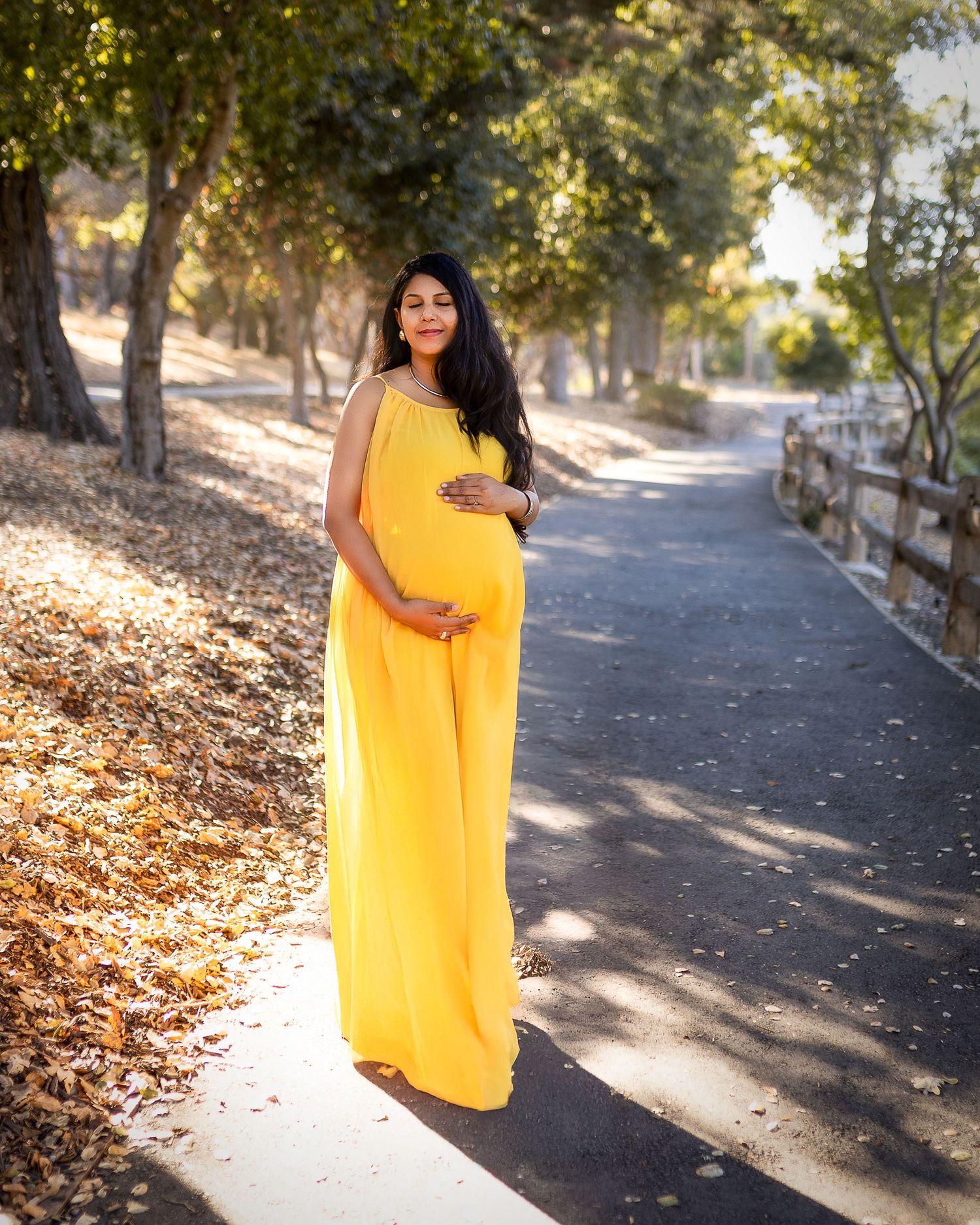 A person wearing a long yellow dress stands on a wooded path with autumn leaves and a rustic fence.