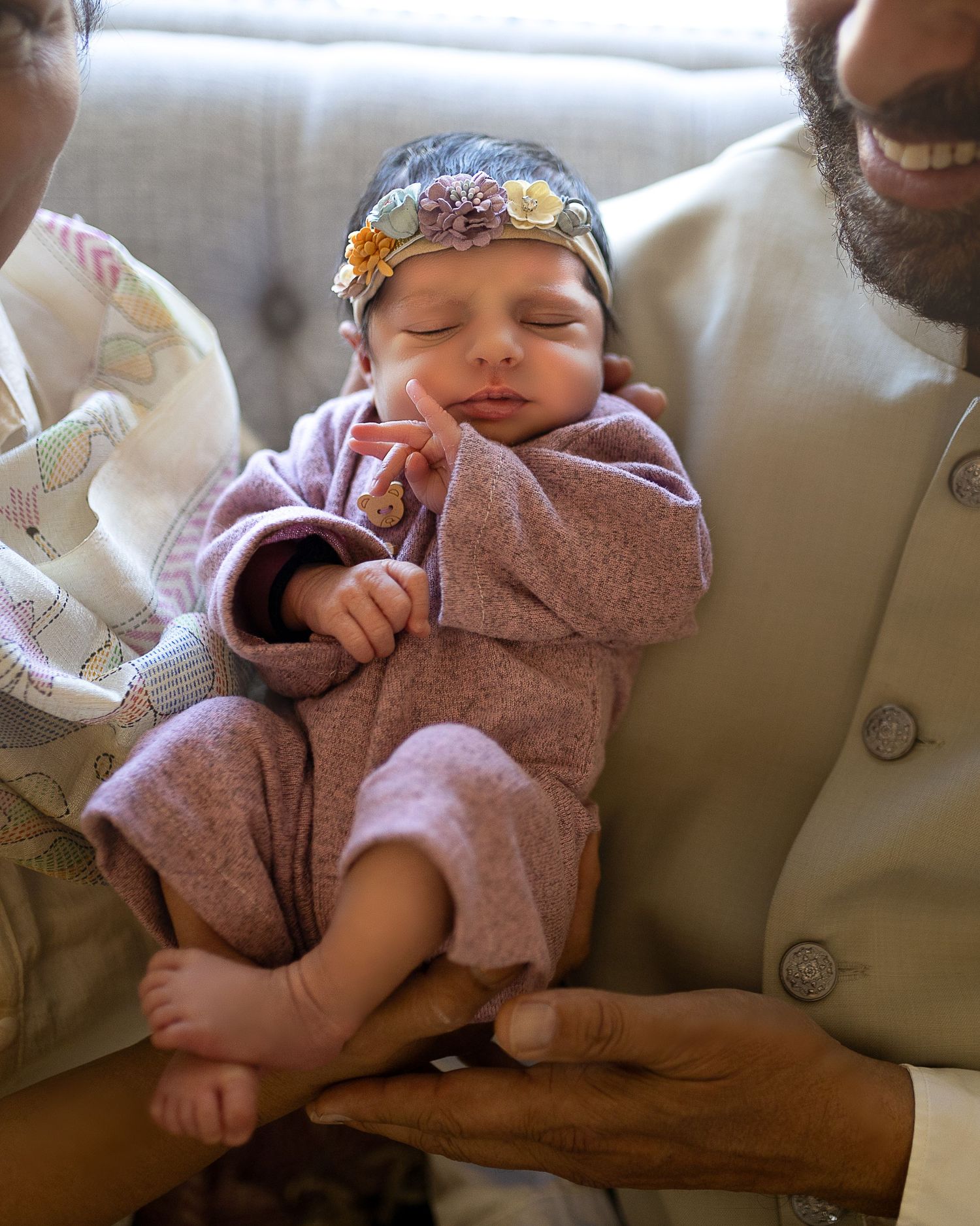A sleeping newborn in a mauve outfit wears a floral headband while being cradled by loving arms.