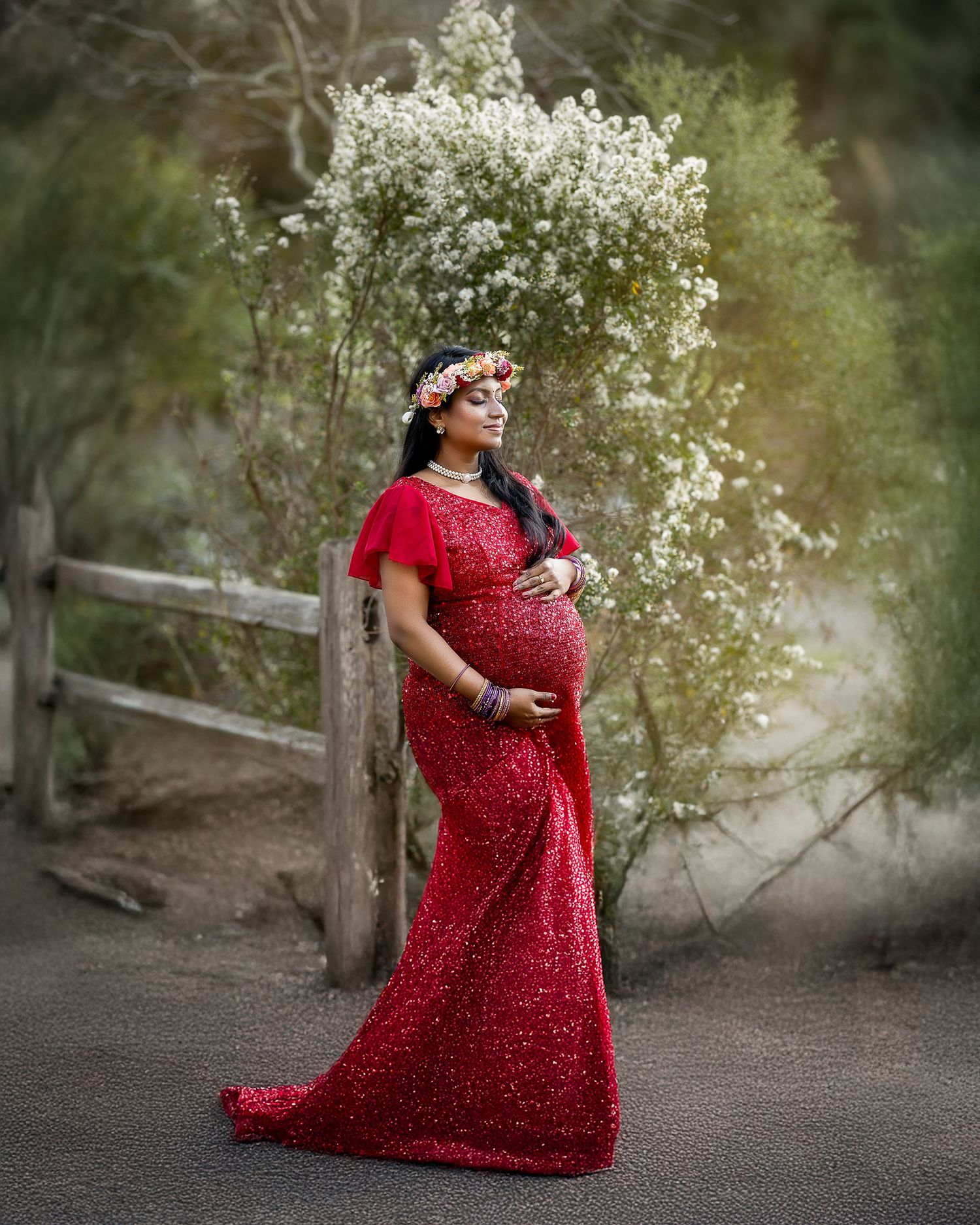 A figure in a flowing red polka dot dress poses gracefully near white flowering bushes and a rustic wooden fence.