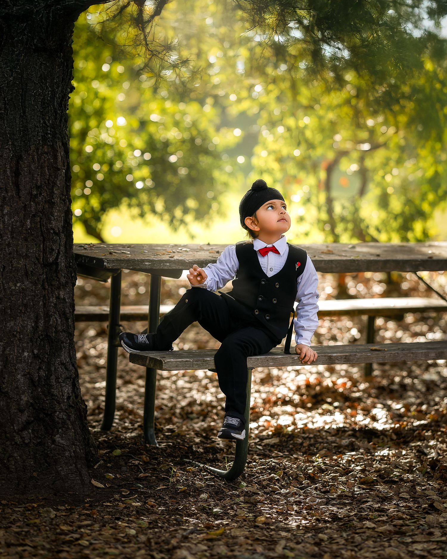 A child in formal attire performs a playful martial arts kick pose against a backdrop of autumn trees and fallen leaves.