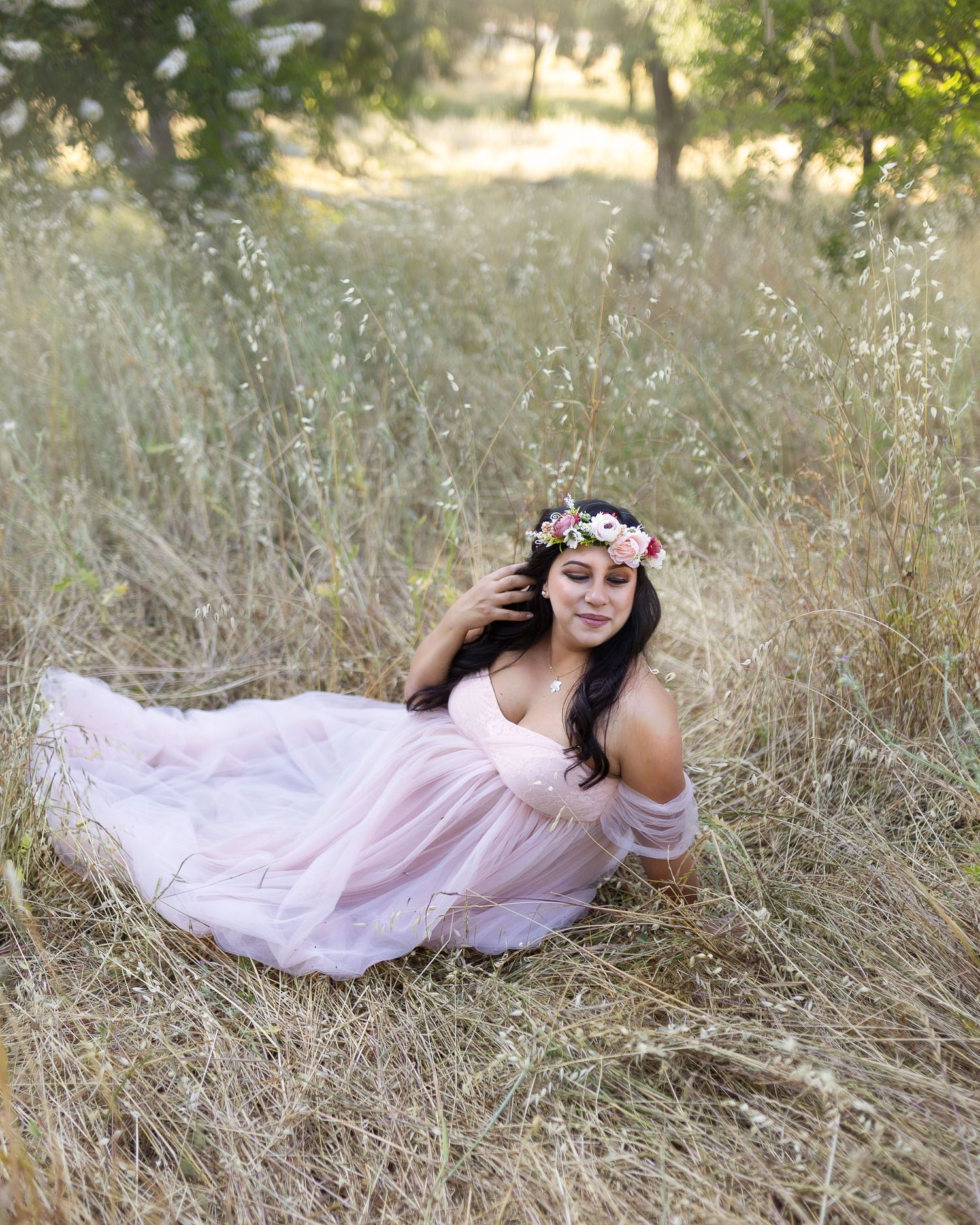 A person in a flowing pink dress lies gracefully in a sun-dappled field of tall dried grass.