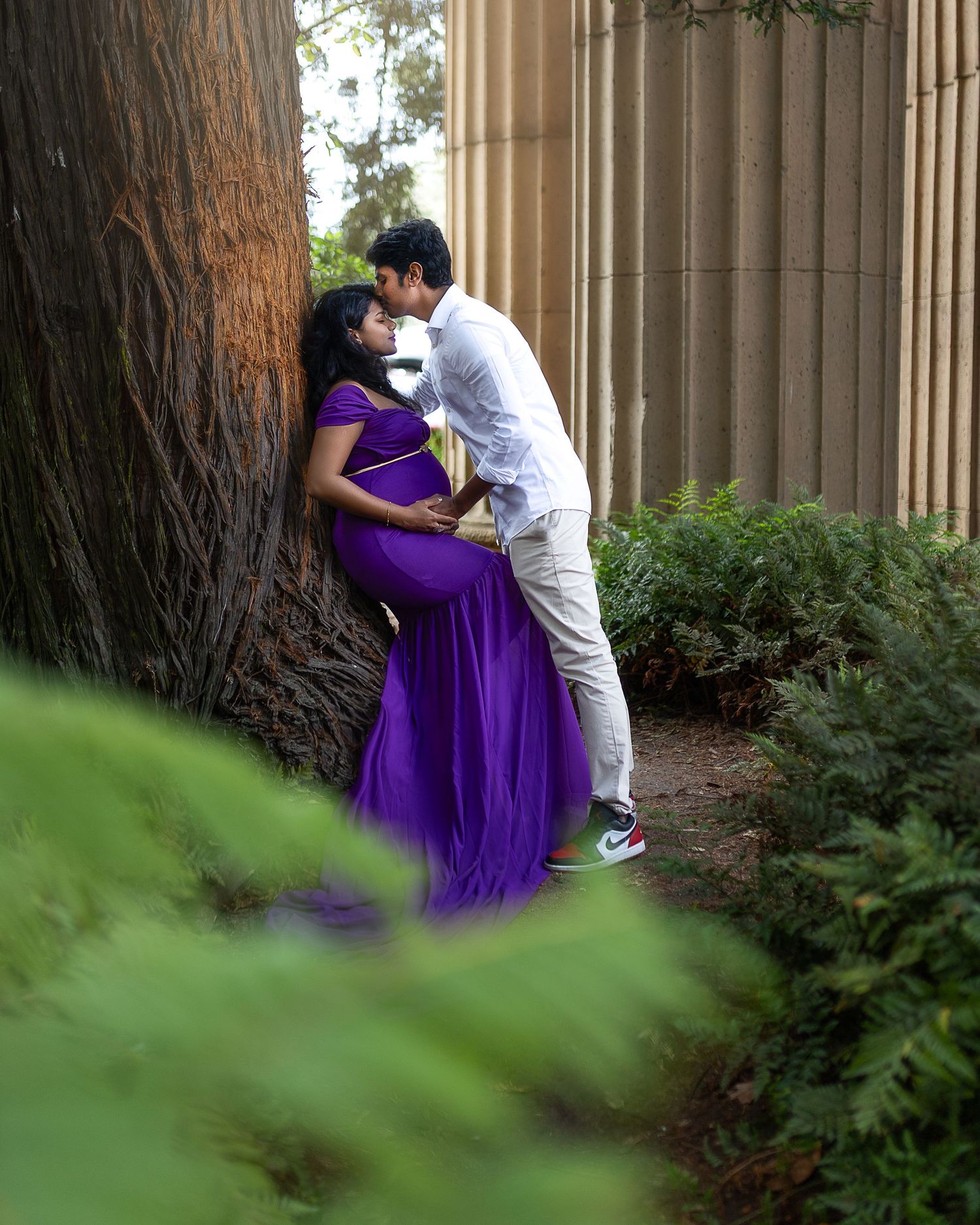 A couple in purple and white attire share an intimate moment against a backdrop of trees and columns.