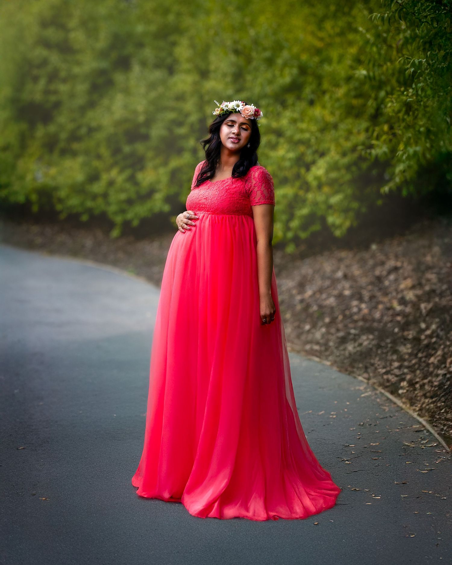 A flowing coral pink tulle maternity gown creates an ethereal silhouette against a wooded outdoor backdrop.
