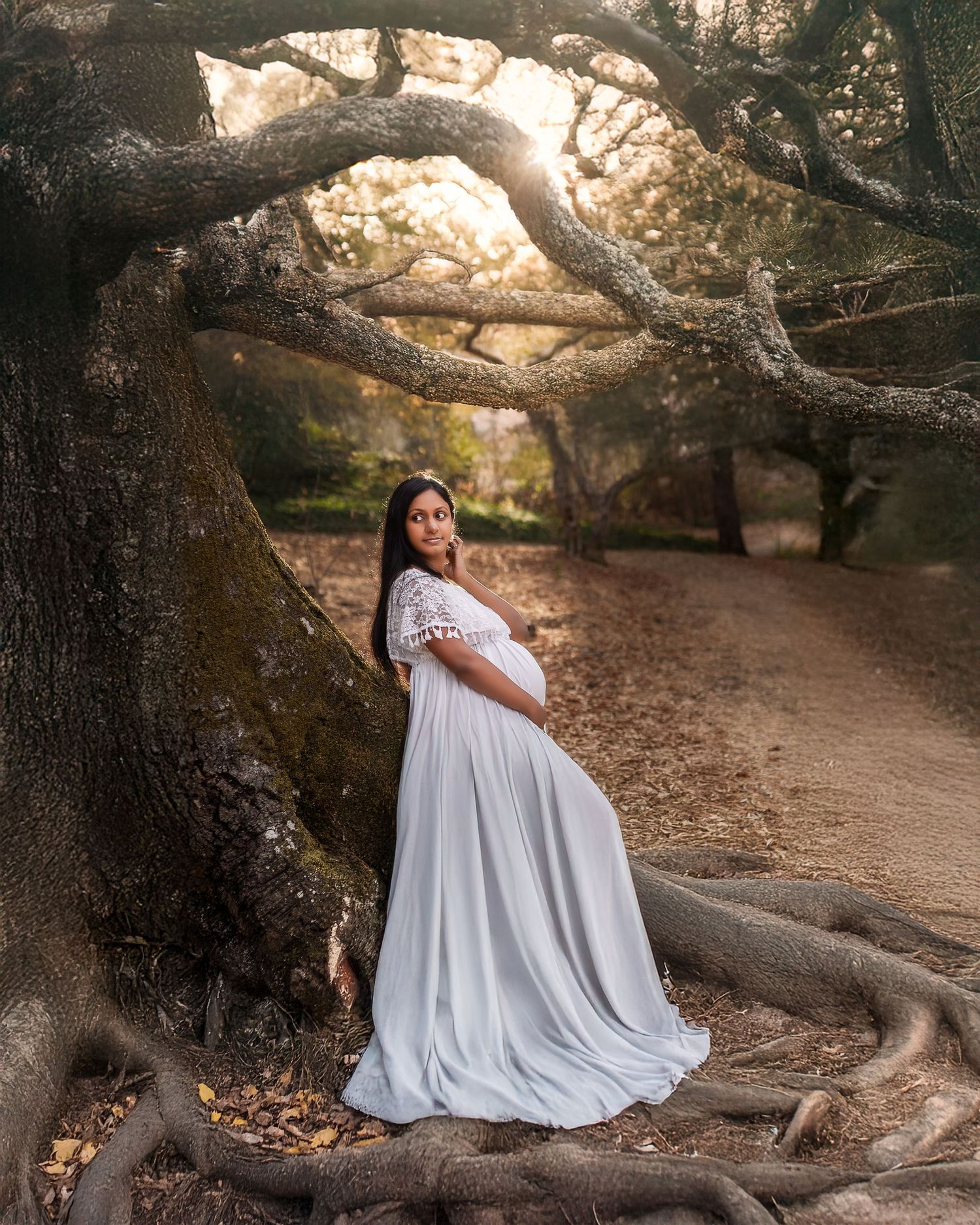 A flowing white dress contrasts beautifully against gnarled tree roots in a dreamy outdoor maternity photoshoot at sunset.
