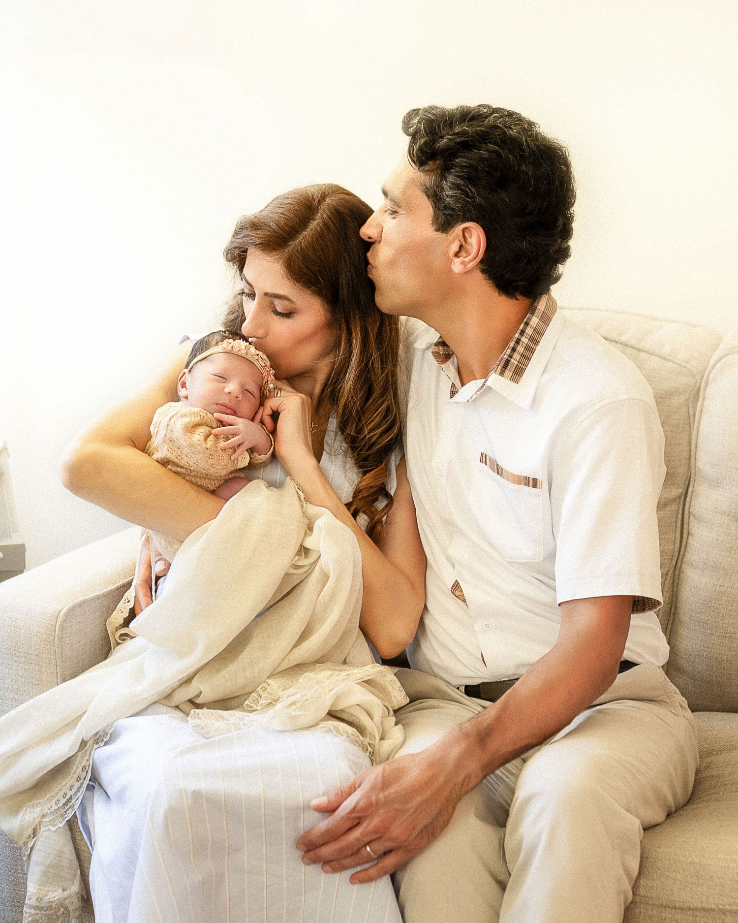 A couple sits on a sofa, both kissing their newborn baby wrapped in a cream blanket.