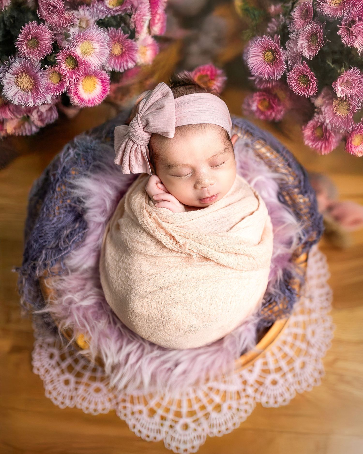 Sleeping newborn wrapped in beige swaddle on lace doily with pink flowers overhead in dreamy photography setup.