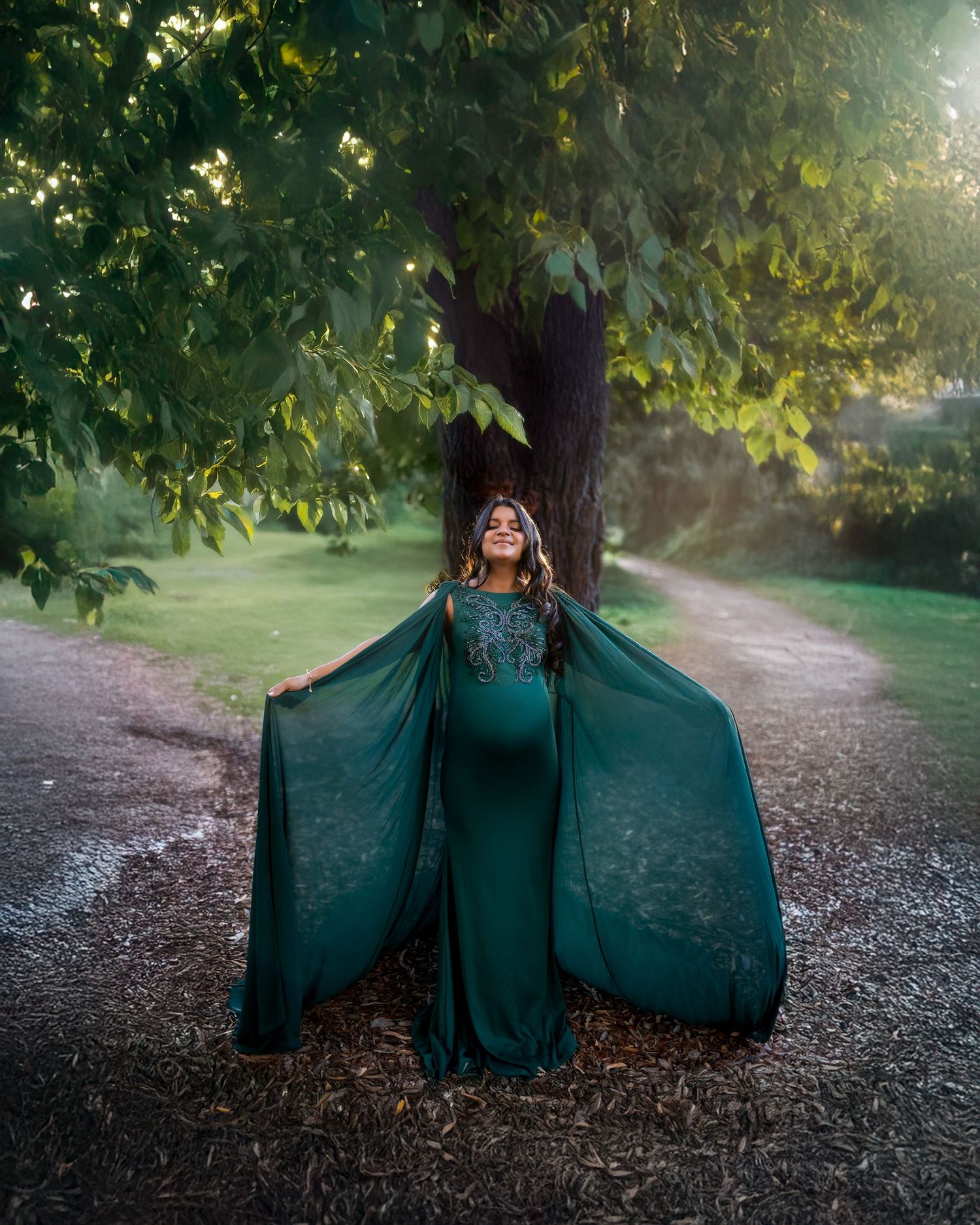 A figure in an elegant emerald green gown and cape stands on a forest path with sunlit trees in the background.