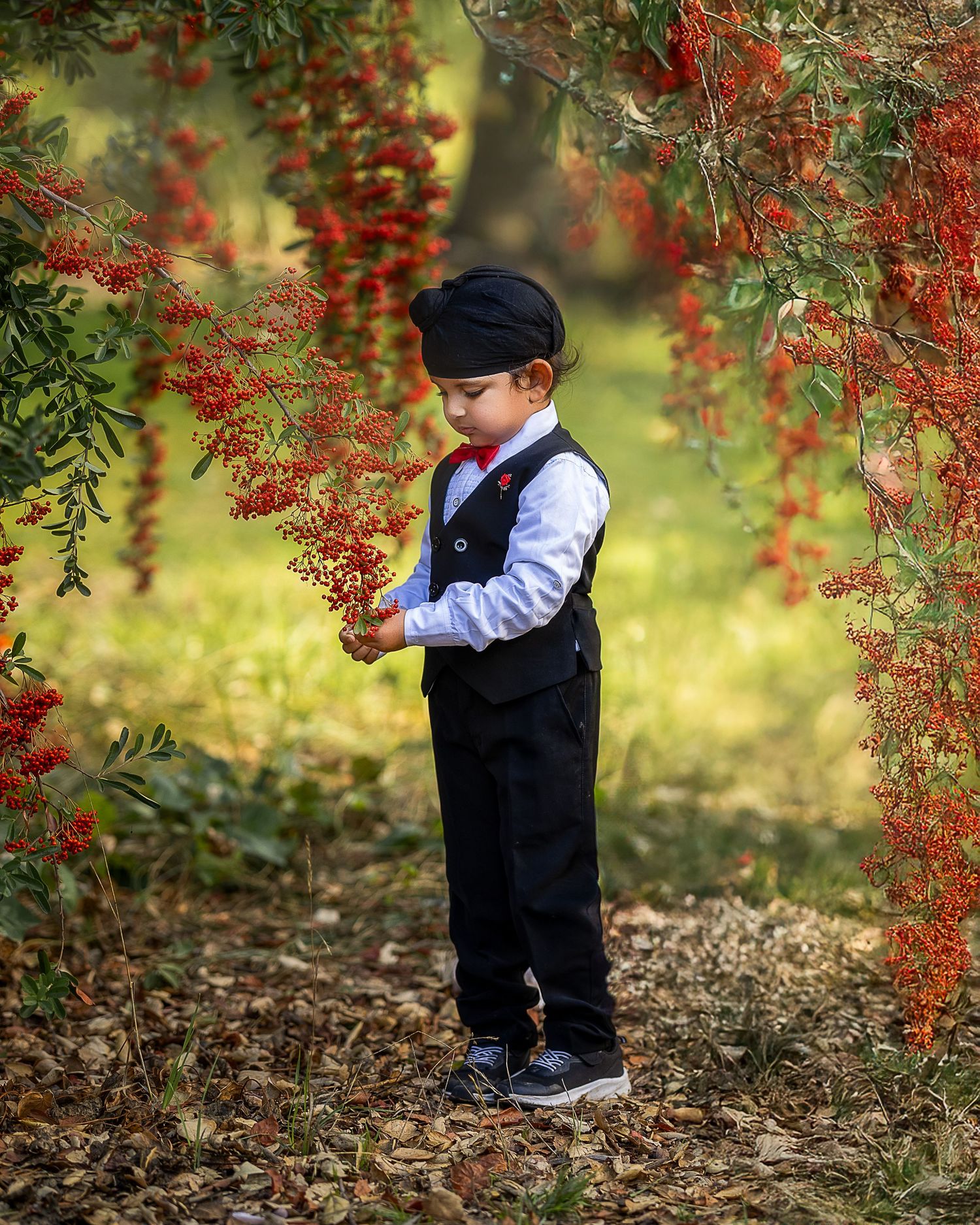 A young child in formal attire stands beneath vibrant red autumn berries against a soft bokeh background.