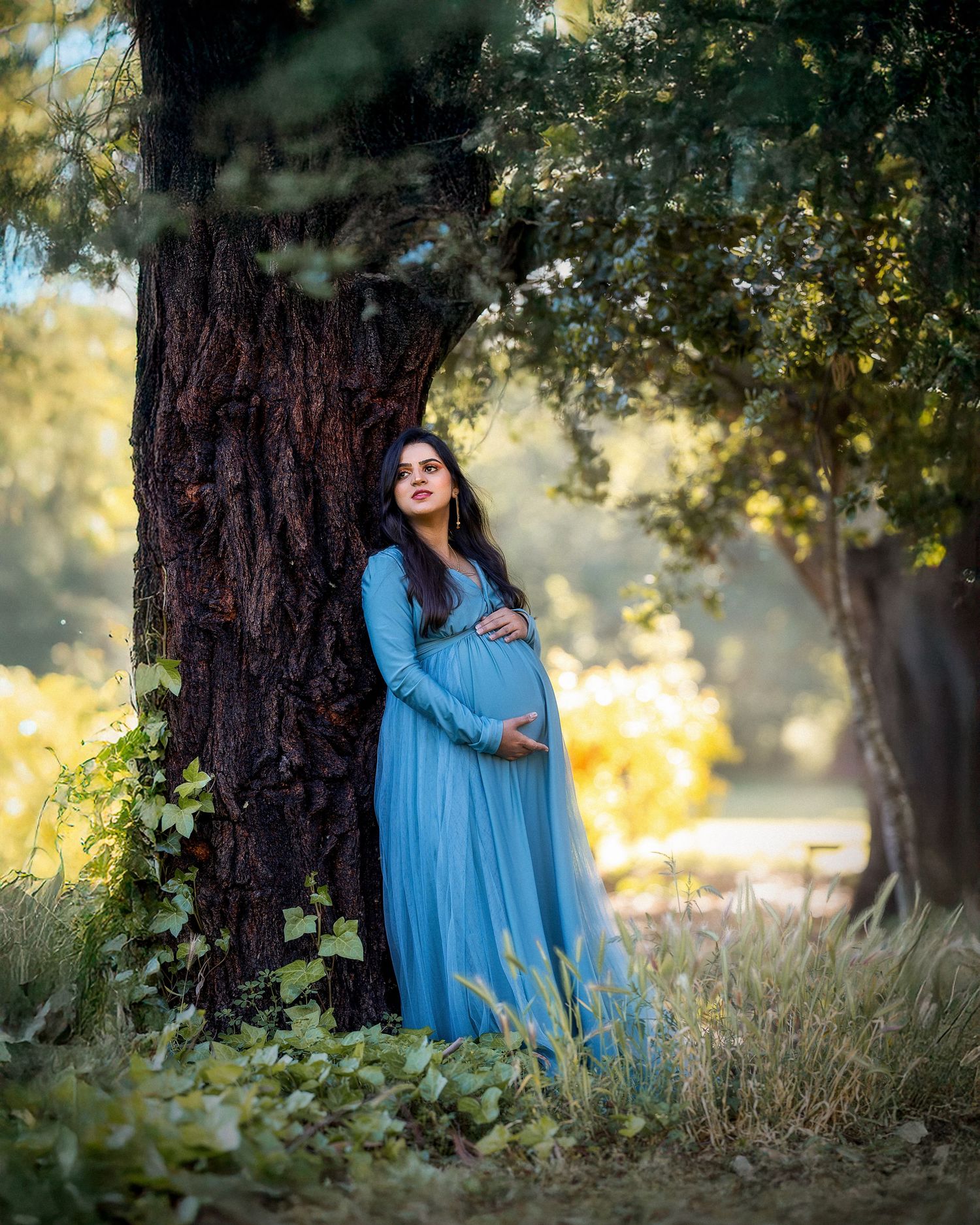 A flowing blue maternity dress contrasts beautifully against a large tree trunk in a dreamy outdoor setting with golden light.