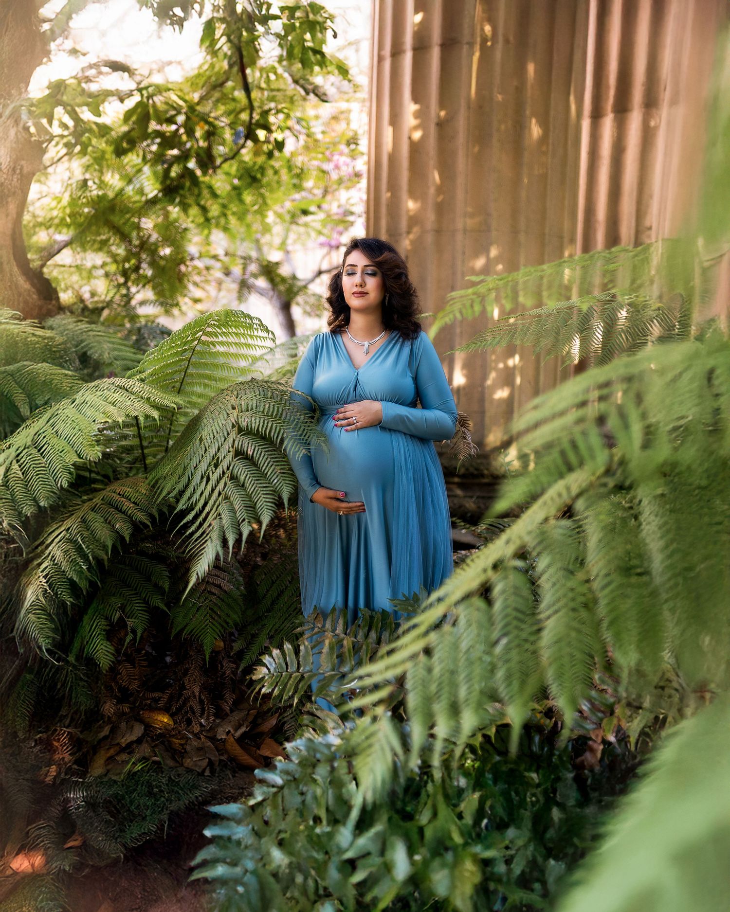 A maternity photo in a lush garden setting with ferns and dappled sunlight, featuring a blue pleated dress.