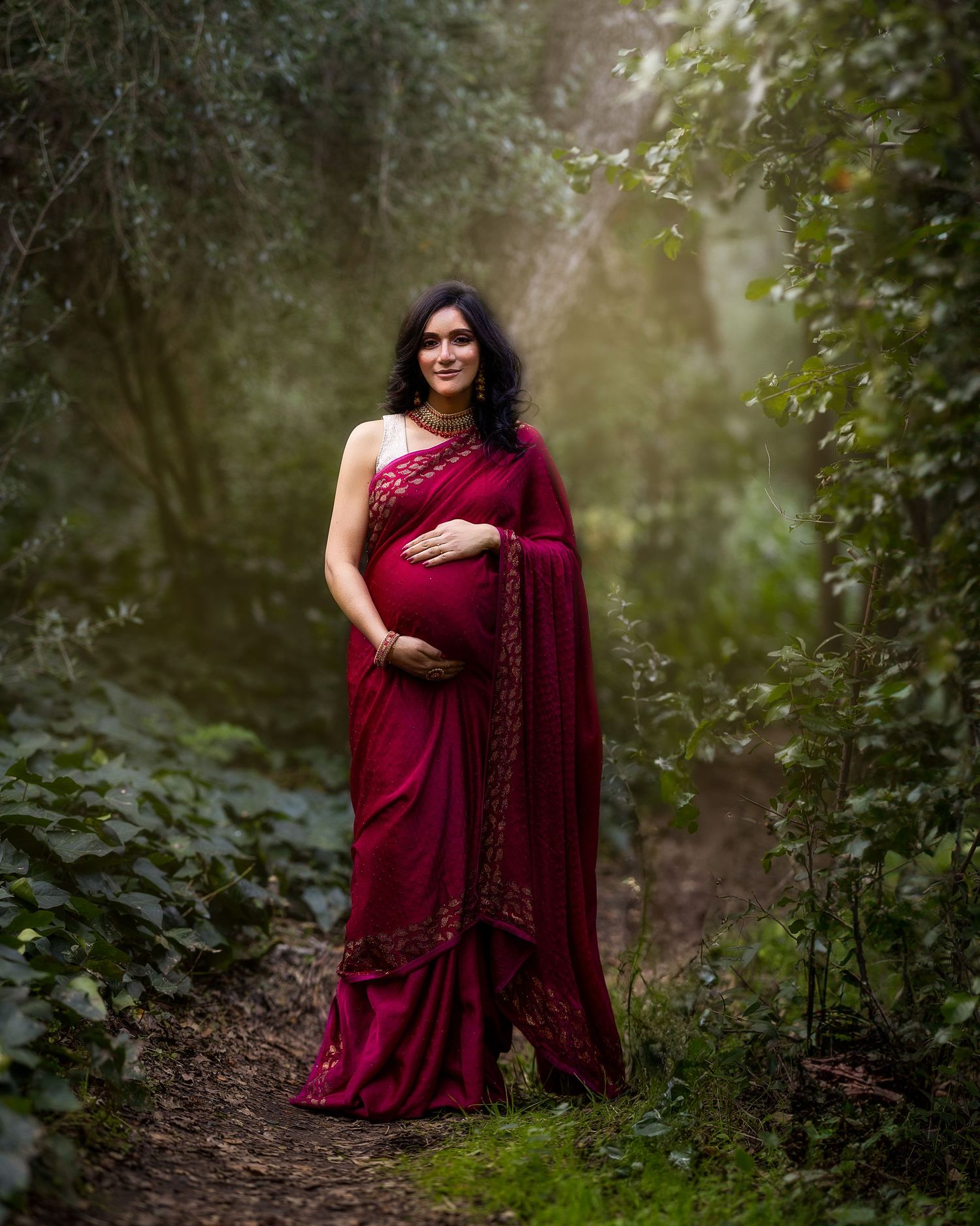 A figure in a deep burgundy dress sits gracefully on a forest path surrounded by lush greenery and dappled sunlight.