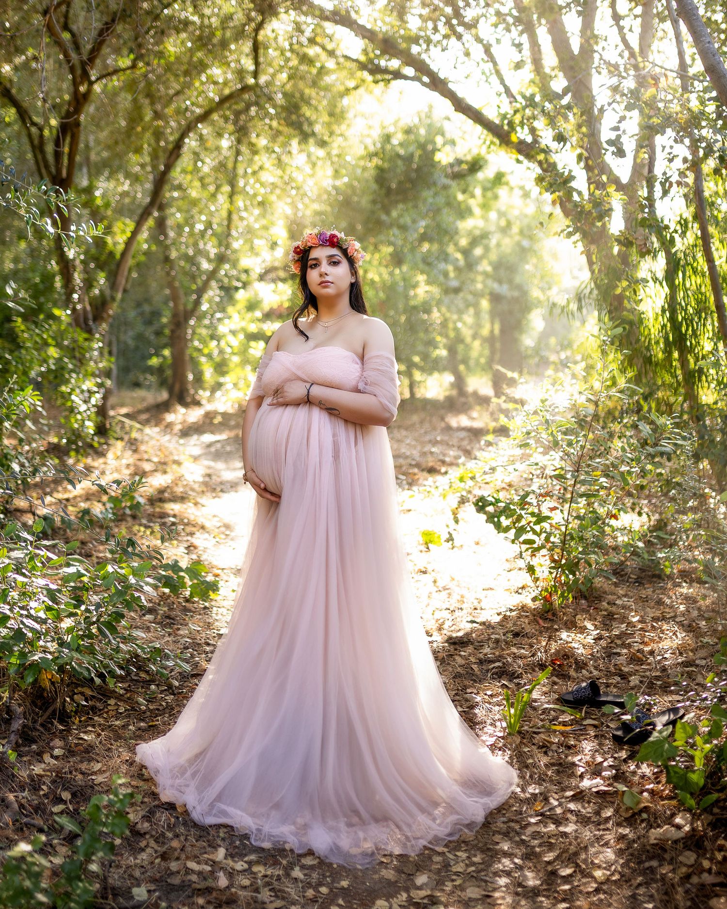 A dreamy maternity photoshoot in a sunlit forest features a flowing pink tulle gown creating an ethereal atmosphere.