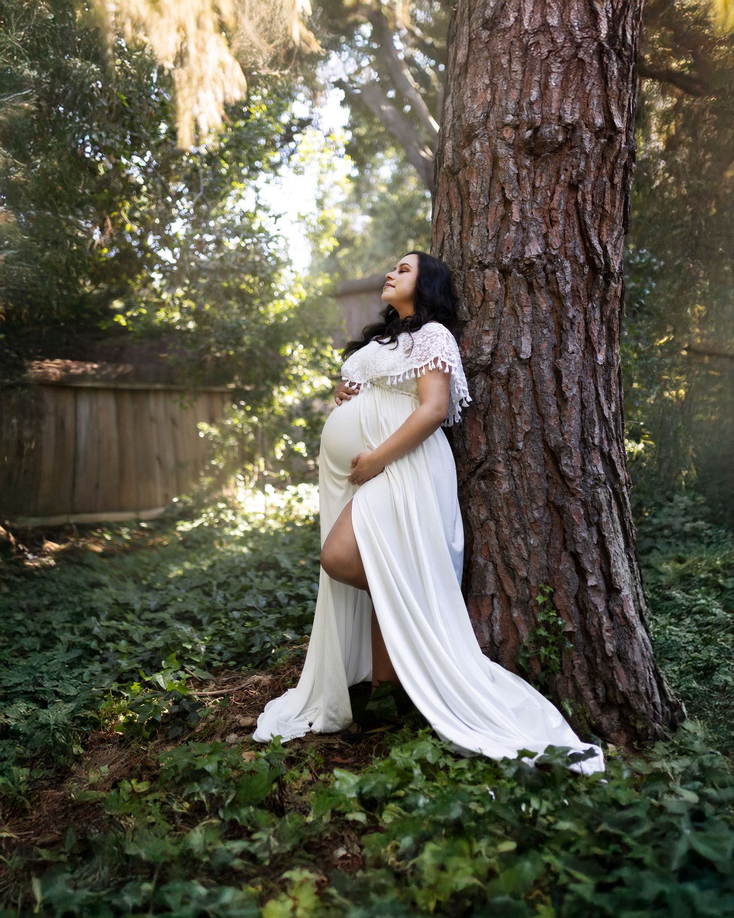 A person in flowing white fabric sits against a large tree trunk in a wooded garden setting with dappled sunlight.