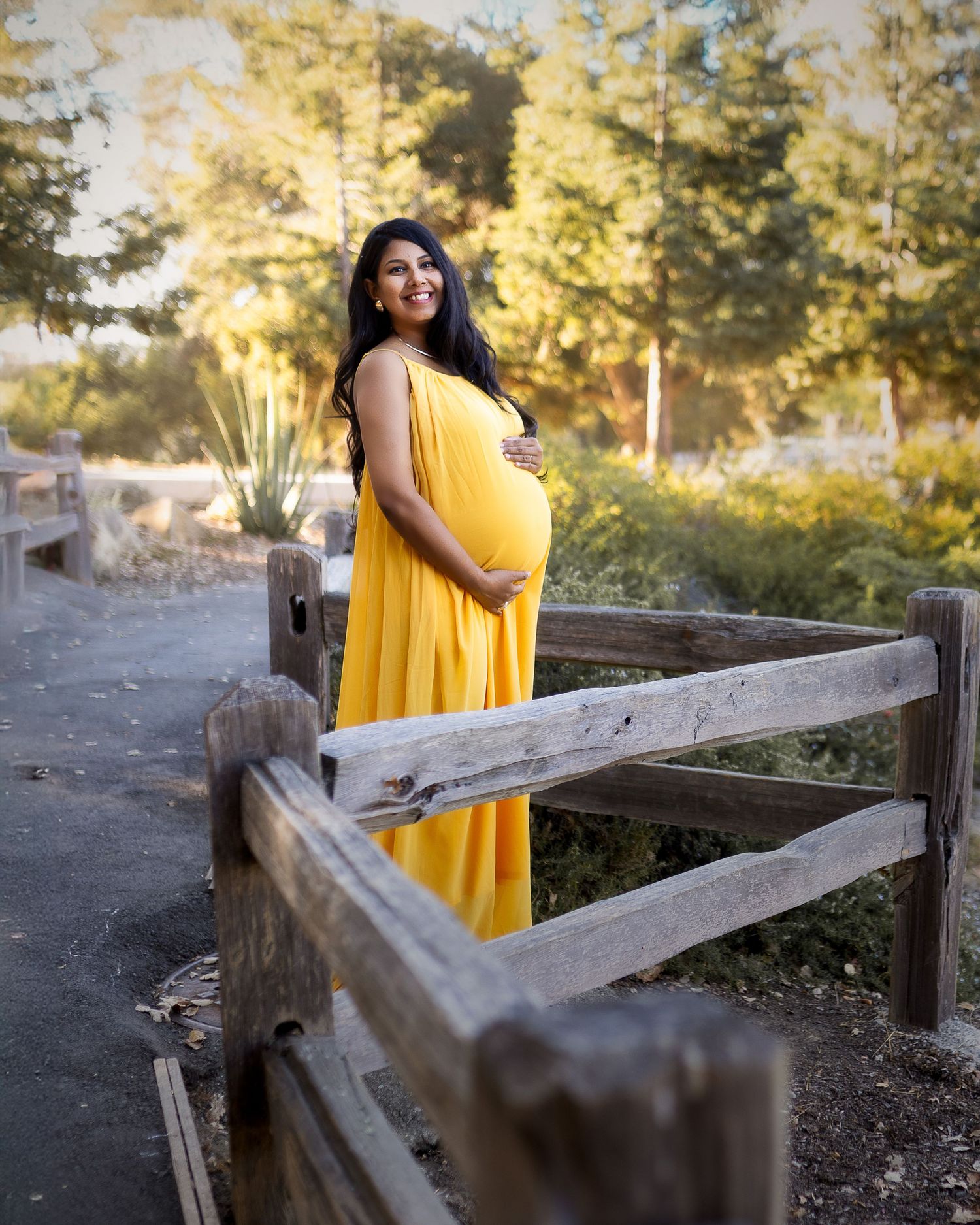 Person in flowing yellow dress poses against rustic wooden fence during golden sunset in outdoor setting.
