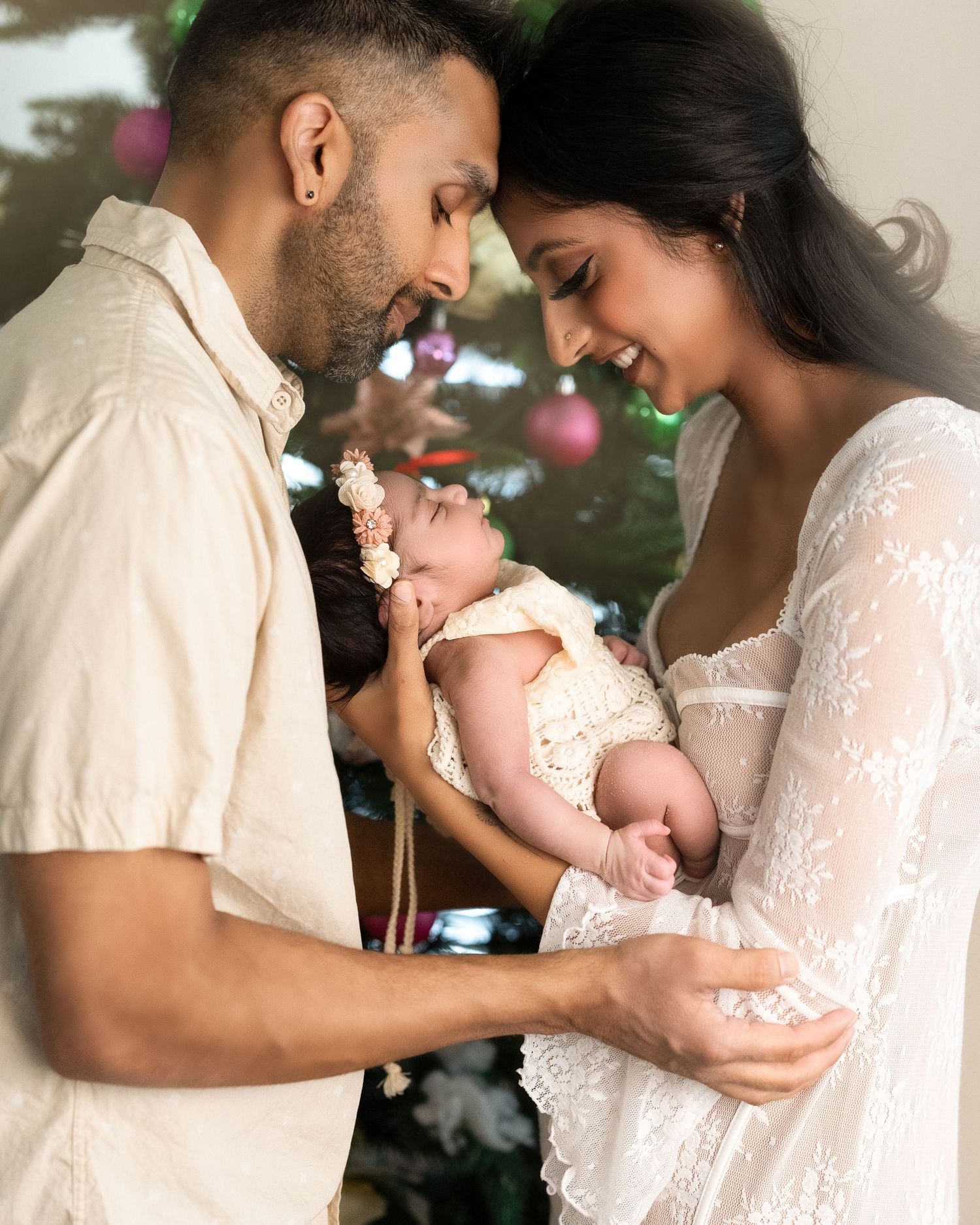 Tender family moment with parents and baby dressed in white lace outfits near Christmas decorations.