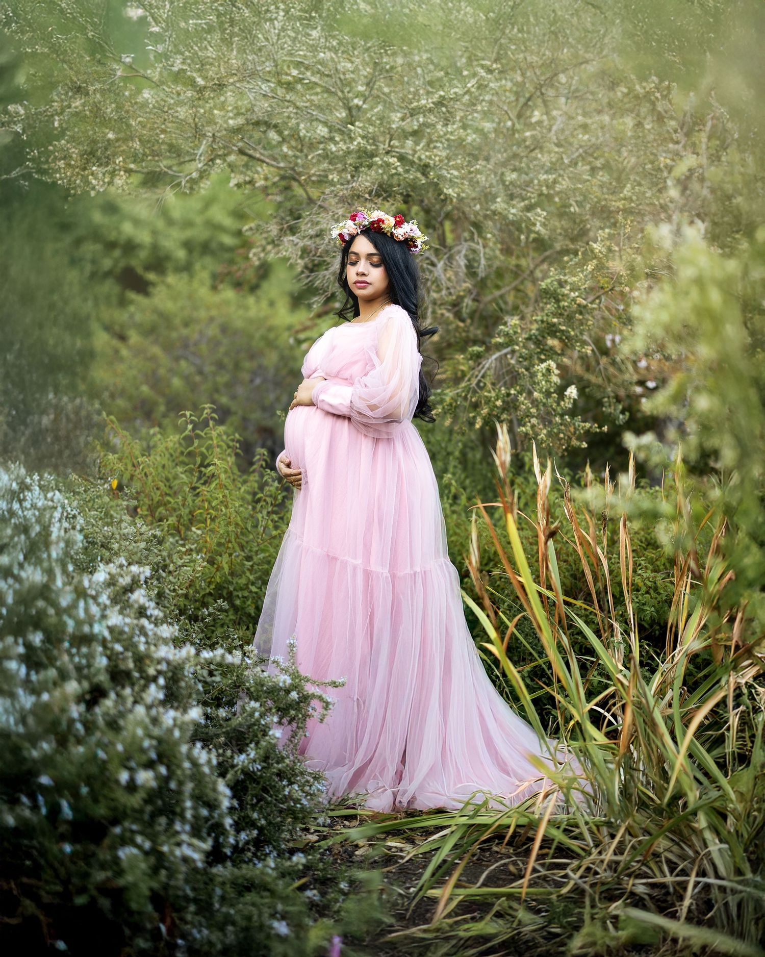 A figure in a flowing pink tulle dress poses gracefully in a garden setting with lush greenery and flowers.