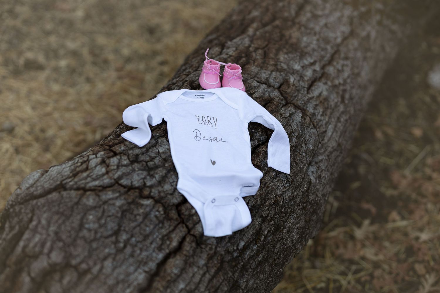 A white baby onesie with pink bow displayed on a tree trunk outdoors.