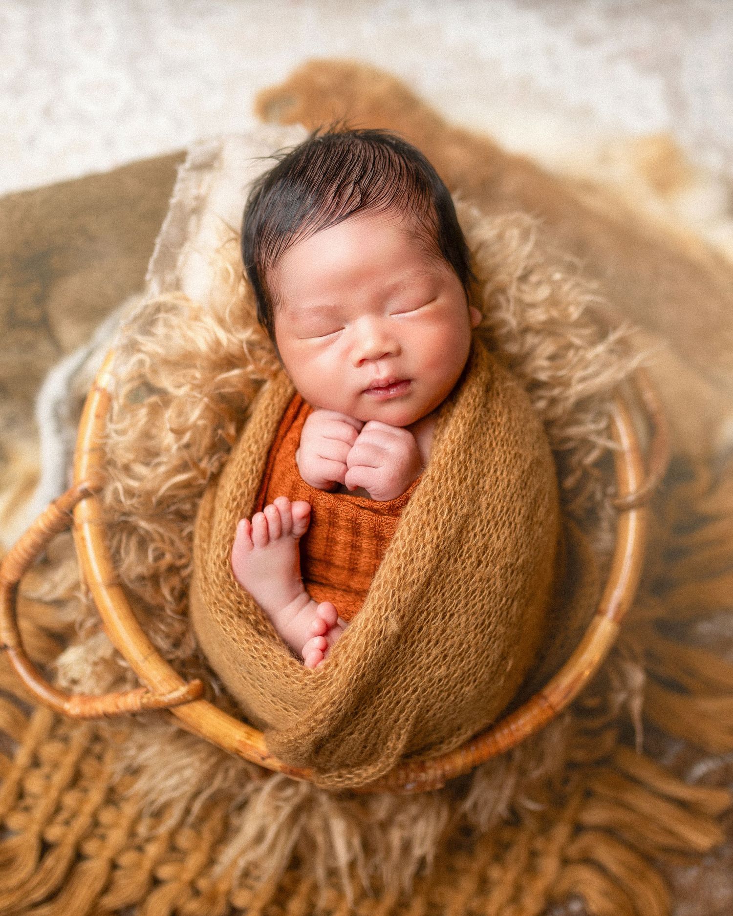 Newborn baby swaddled in orange knit wrap sleeping peacefully in a wicker basket with fluffy tan fur lining.