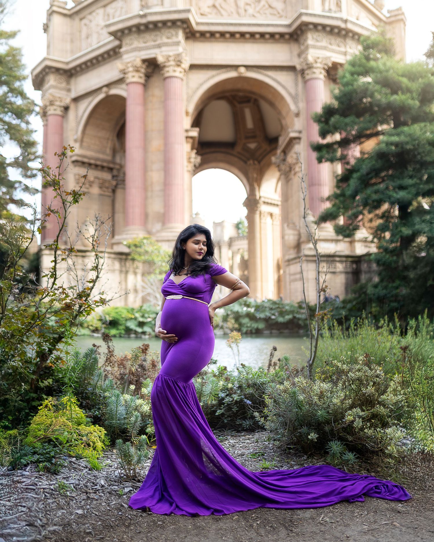 A person in a flowing purple maternity gown poses gracefully in a garden setting with classical architecture.