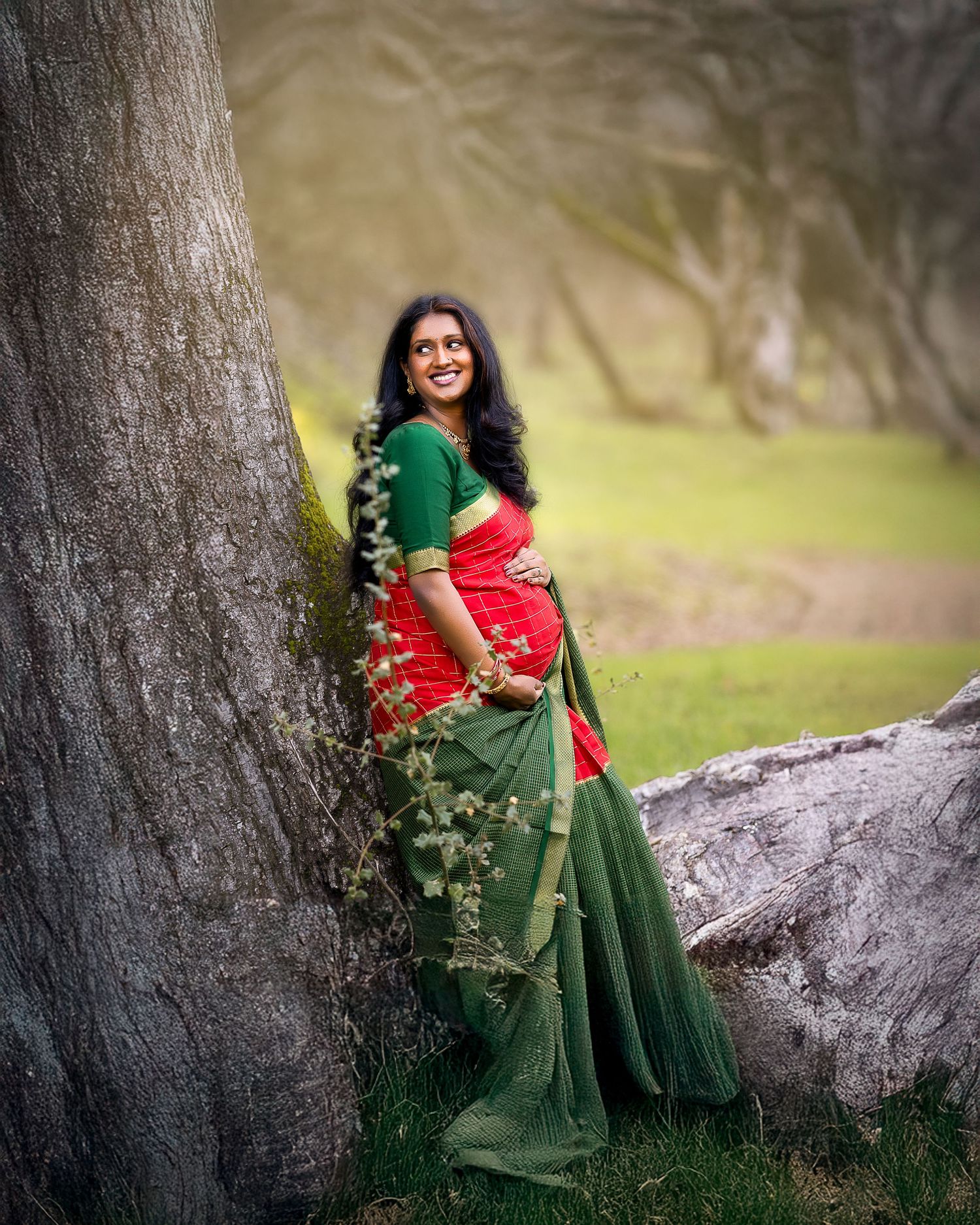A woman in a red and green saree poses gracefully against a tree trunk in a natural outdoor setting.