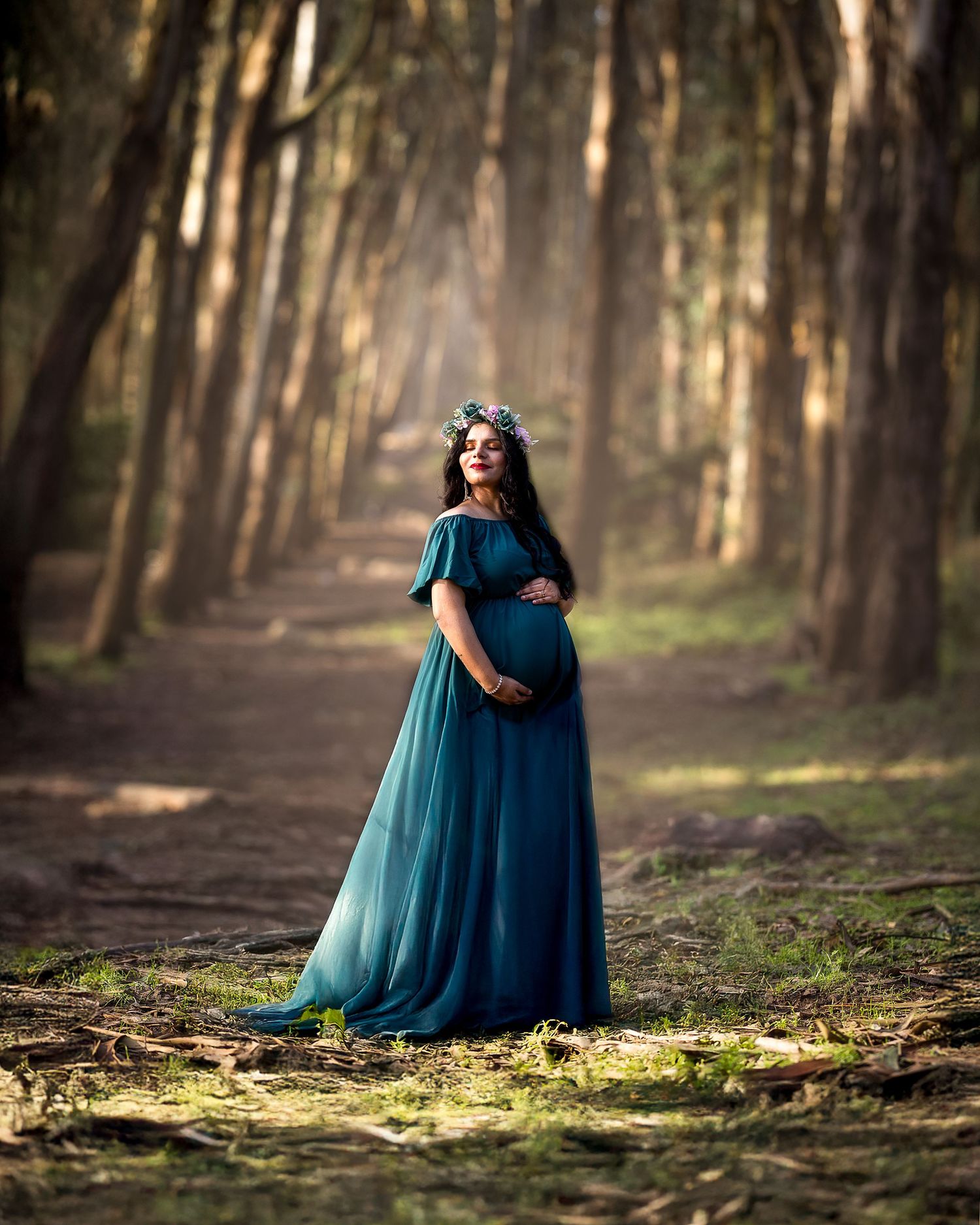 A figure in a flowing teal maternity gown stands on a forest path surrounded by tall trees in golden sunlight.
