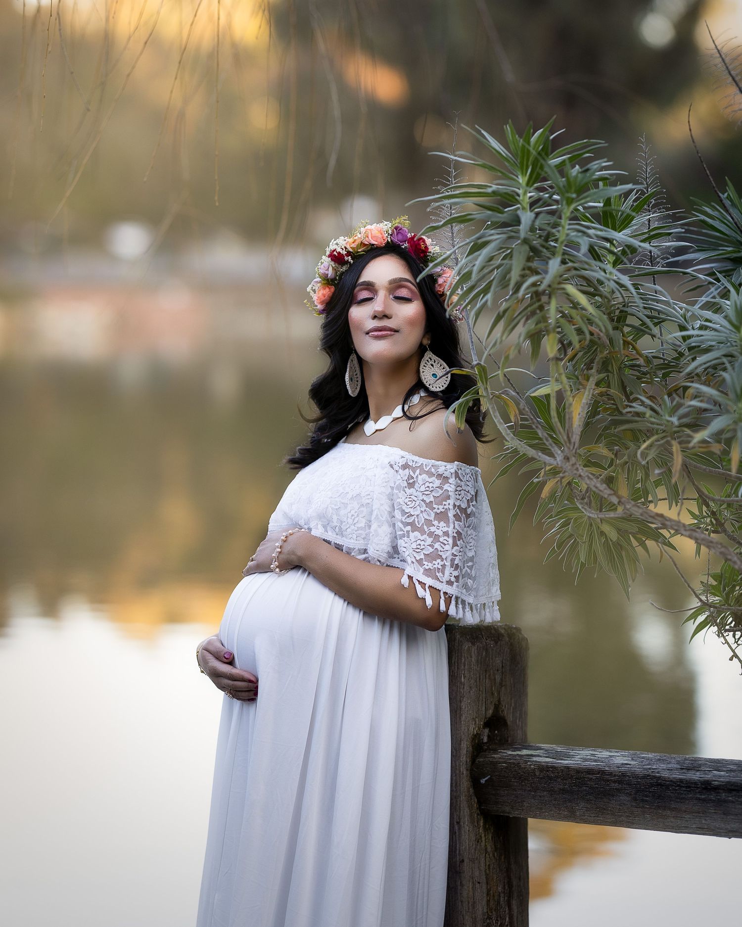 A pregnant woman in a flowing white dress stands by a lake with autumn foliage in the background.