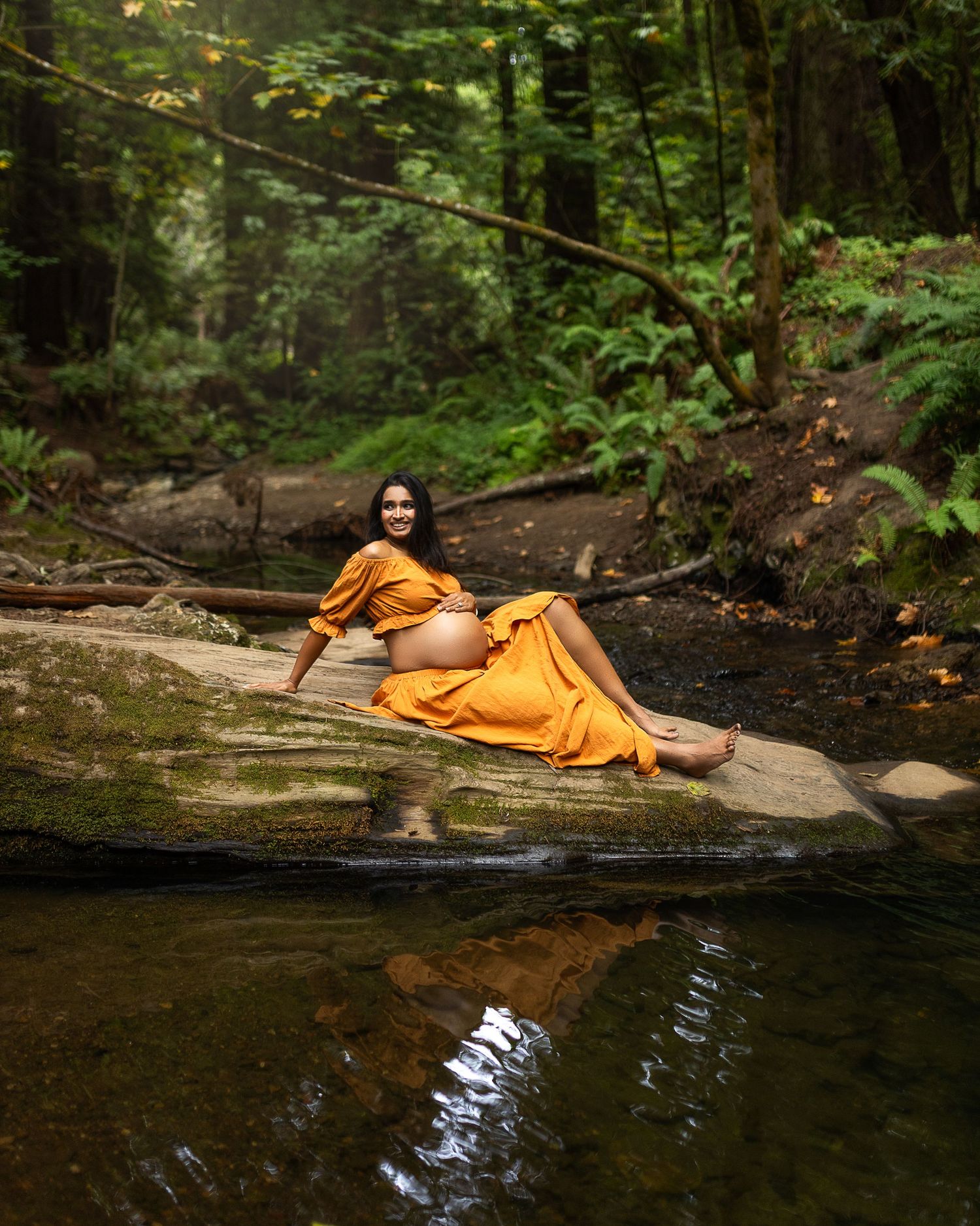 Someone in an orange robe relaxes on rocks by a tranquil forest stream on a peaceful summer day.