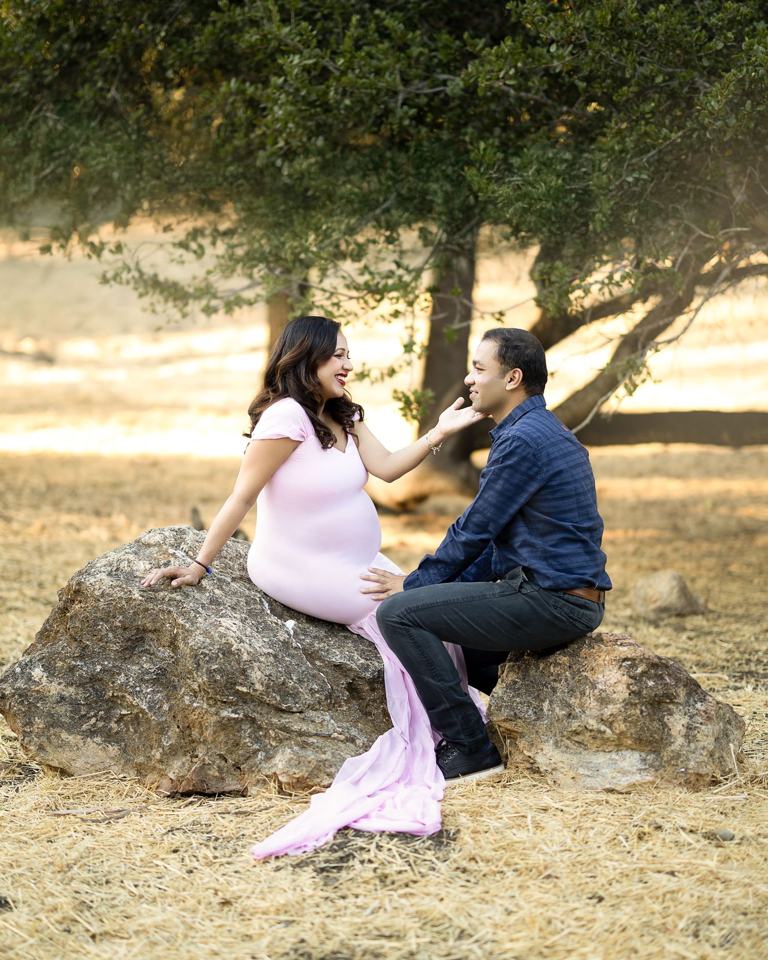 A couple in pink and blue outfits sits together on a large boulder in a sunlit outdoor setting.