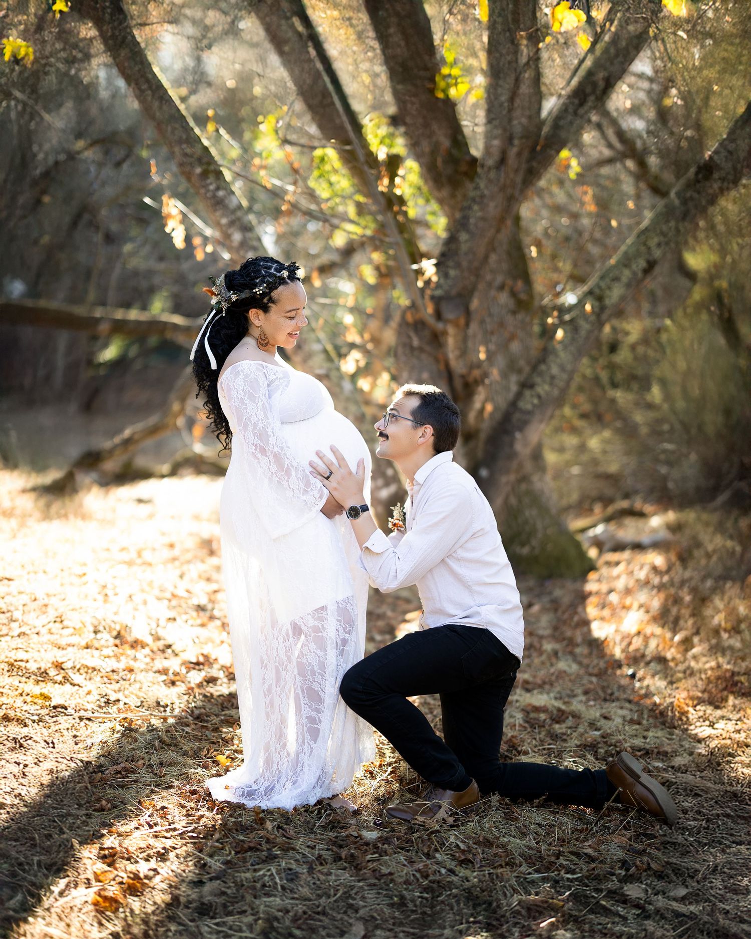 A romantic couple in white clothing poses together under autumn trees in a dreamy outdoor photoshoot setting.