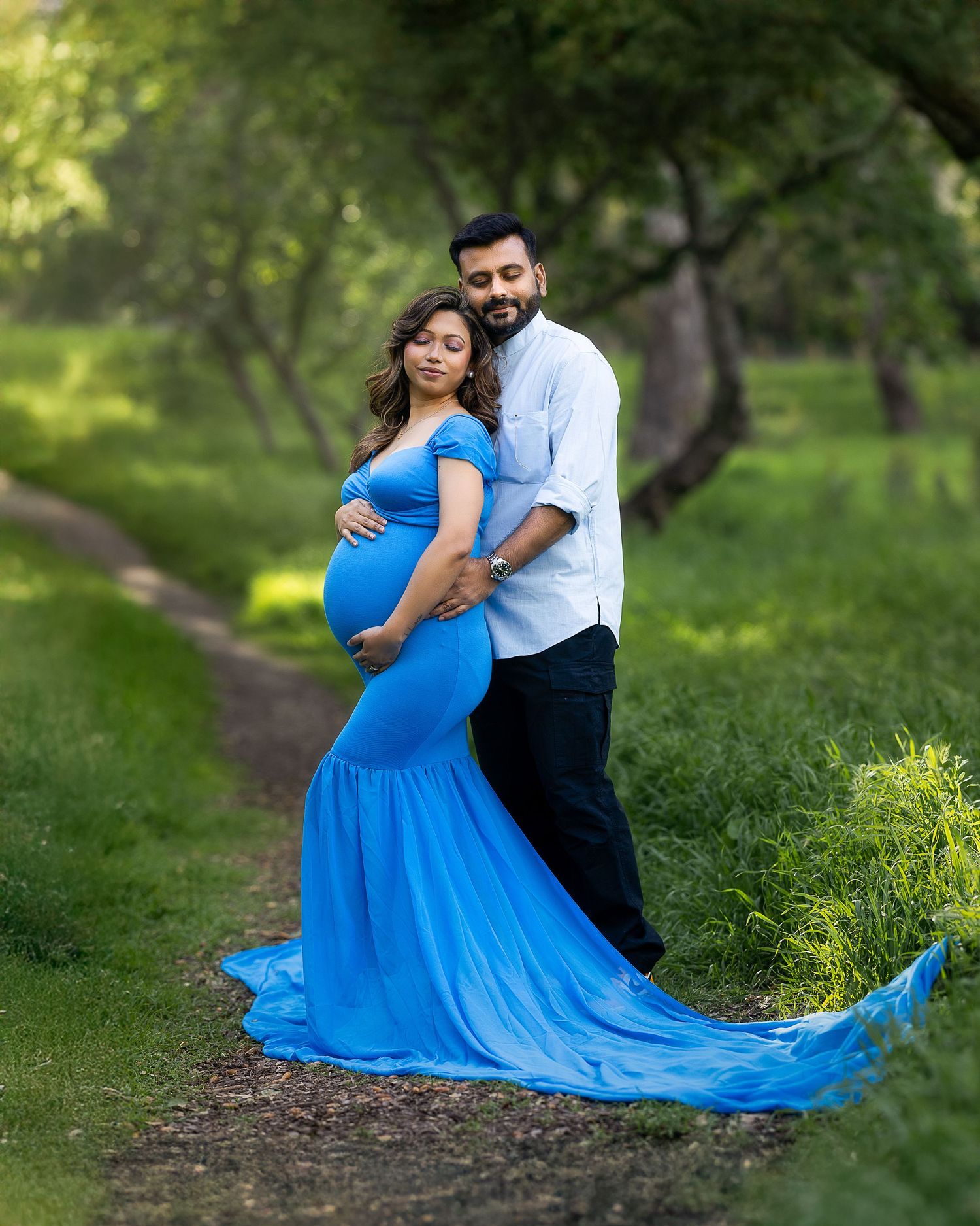 A couple in a romantic outdoor photoshoot, with the woman wearing a flowing bright blue dress against a lush green backdrop.