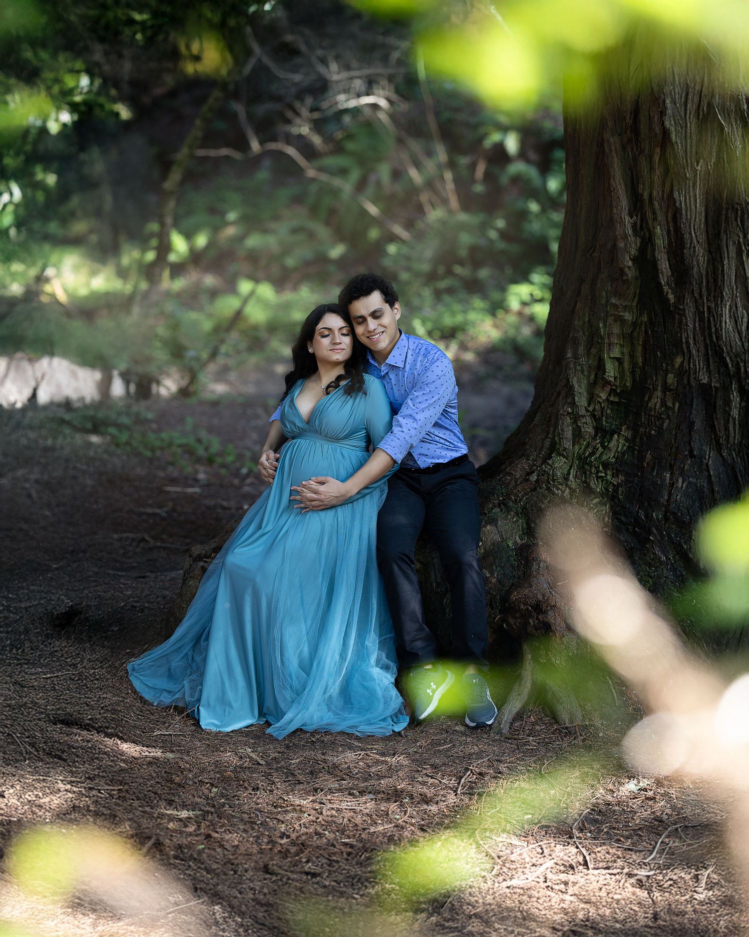 A couple poses for a maternity photo under a large tree in a forest setting, with the woman wearing a flowing blue dress.