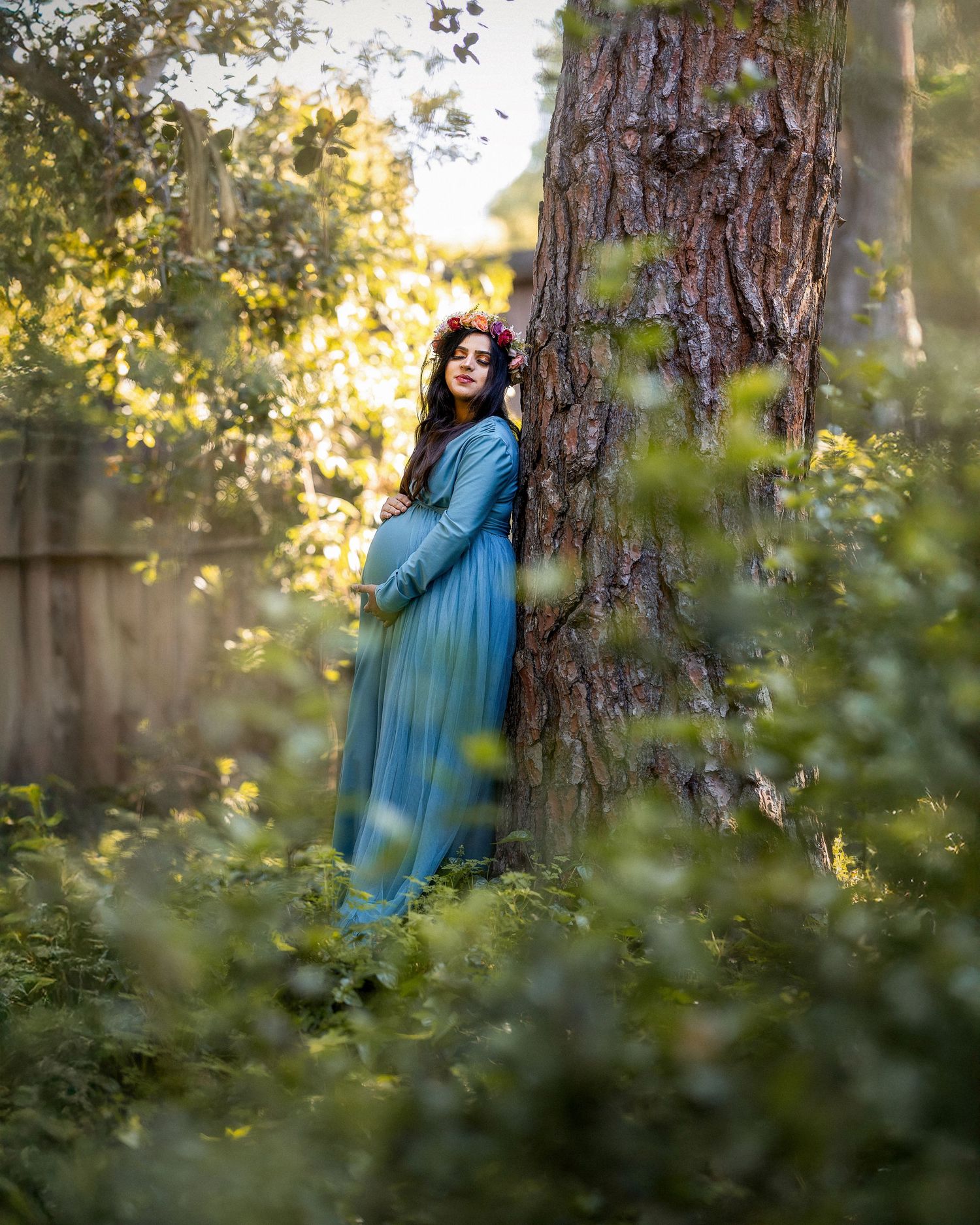 A person in a flowing blue dress poses dreamily against a large tree trunk in a sunlit forest setting.