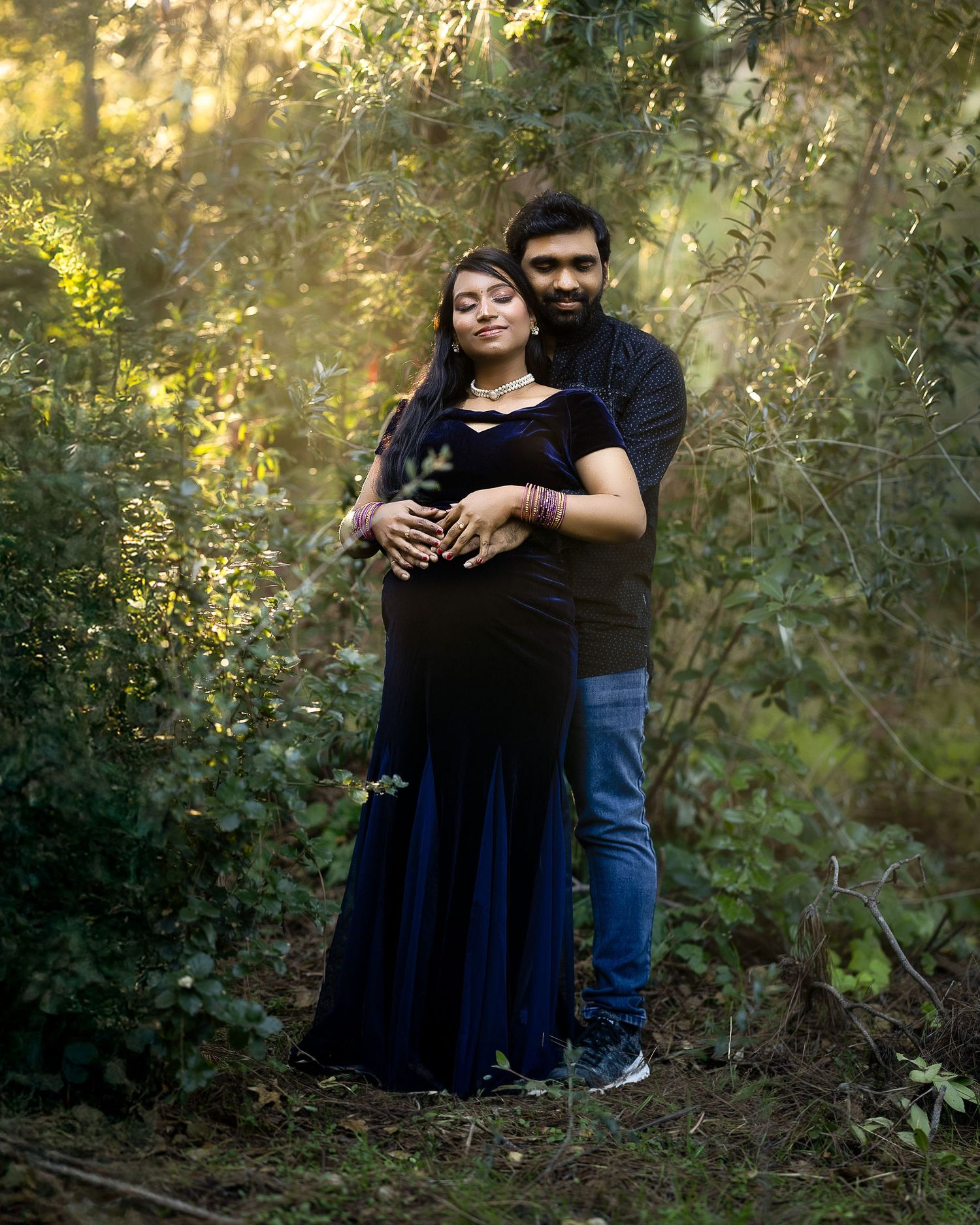 A couple embraces in a romantic outdoor photoshoot among lush greenery with golden sunlight filtering through the trees.