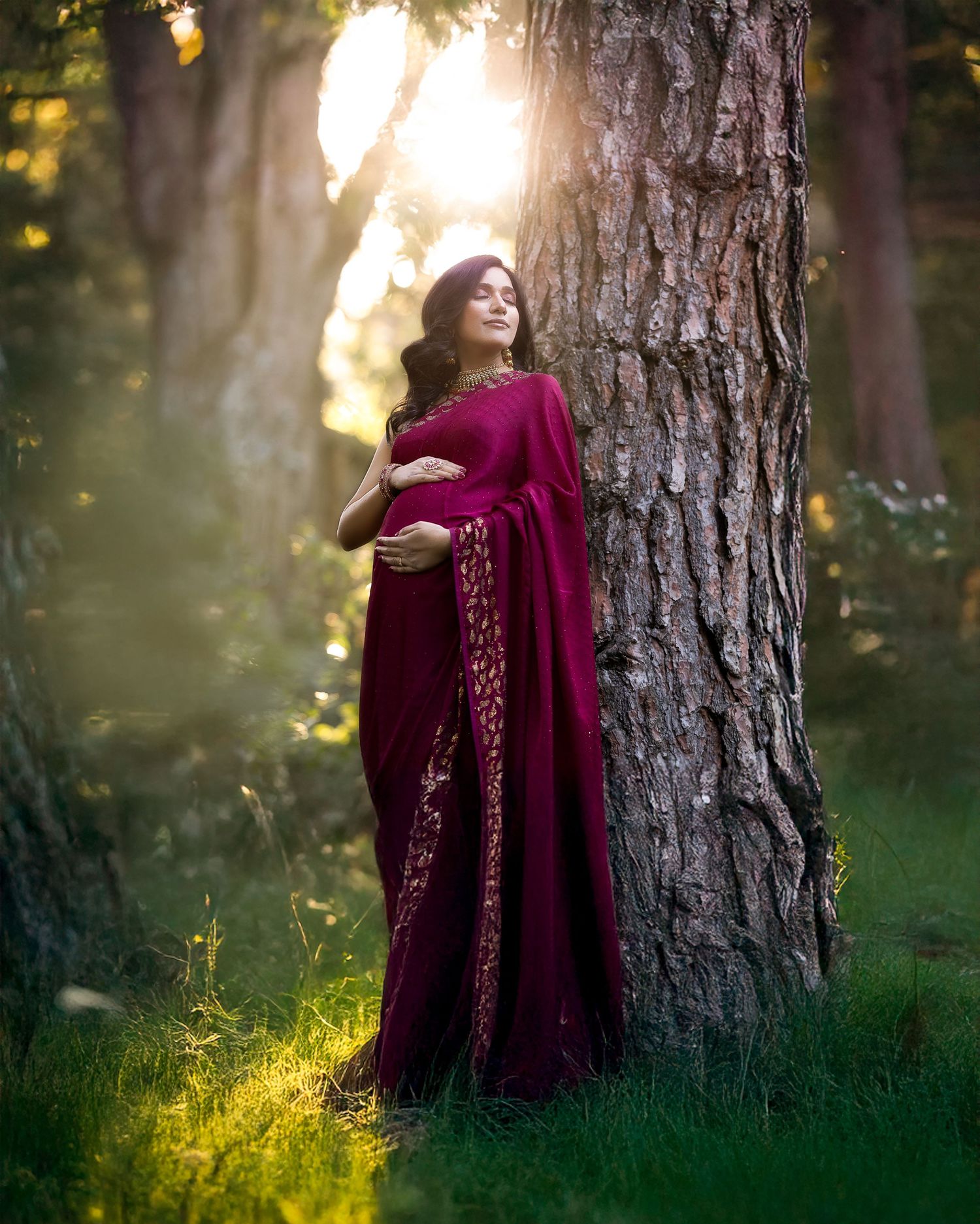 Person in burgundy dress stands dreamily by a large tree trunk in a sunlit forest setting during golden hour.
