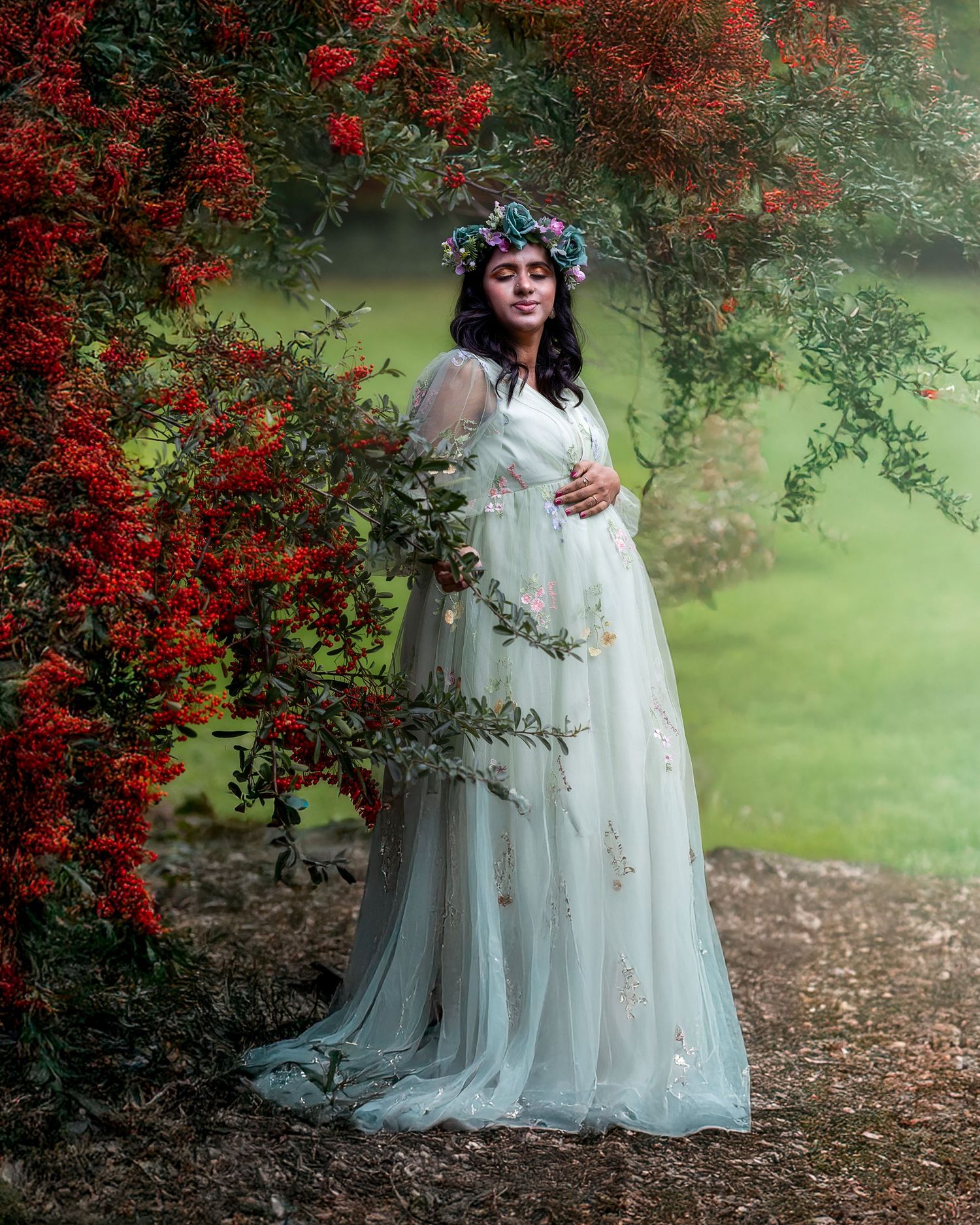 A flowing white maternity gown is captured in a dreamy garden setting with vibrant red flowering bushes.
