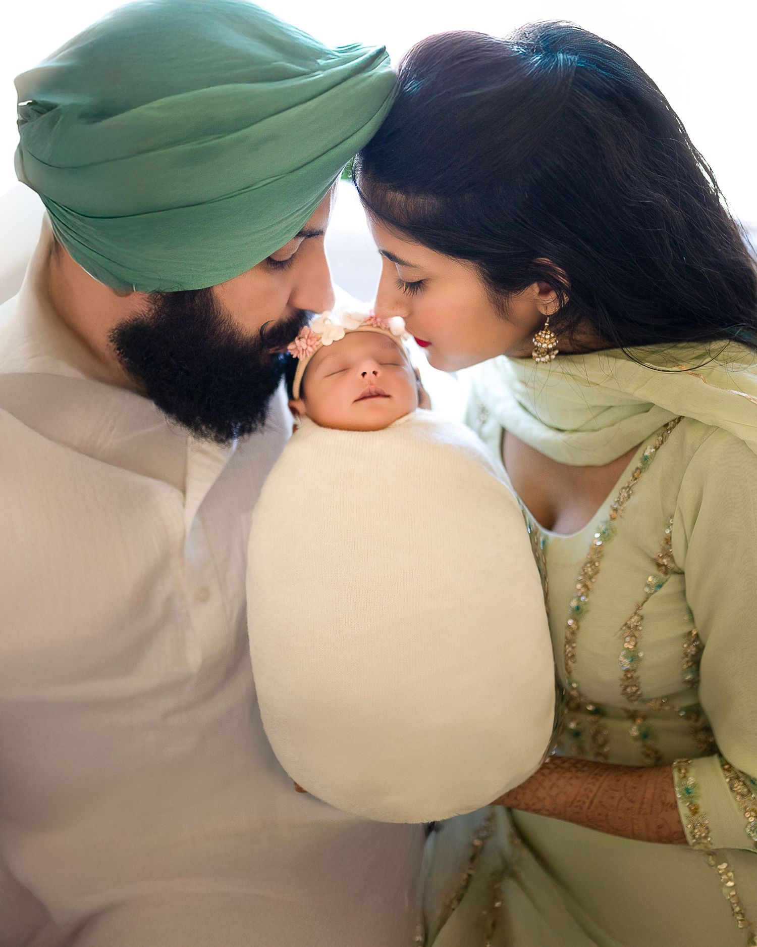 A family shares an intimate moment with their newborn baby wrapped in white, wearing traditional Indian attire.