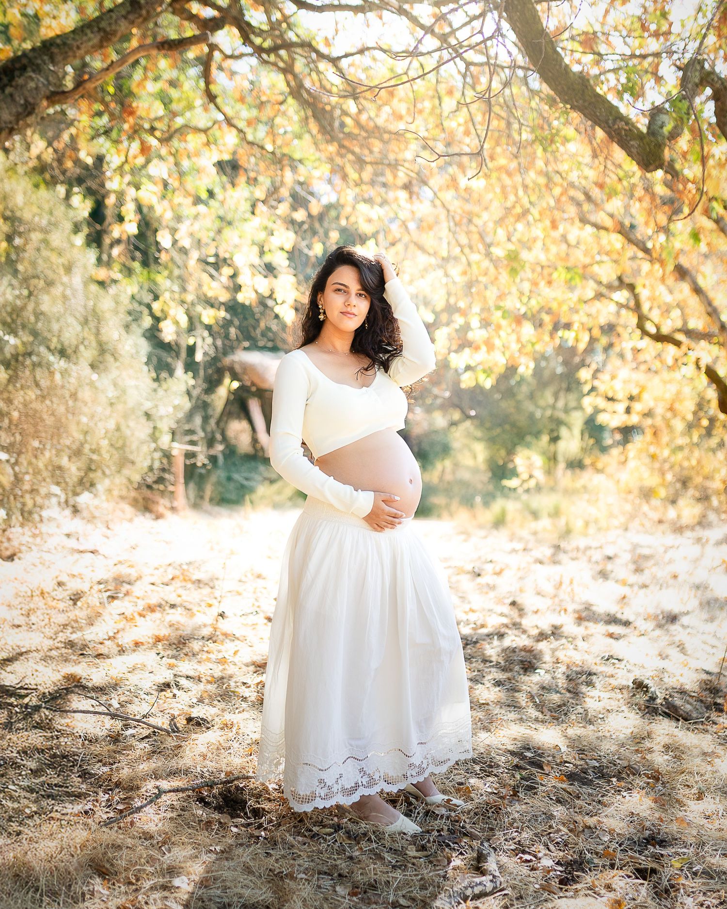 A person in a flowing white lace dress poses gracefully among autumn leaves in a sunlit outdoor setting.