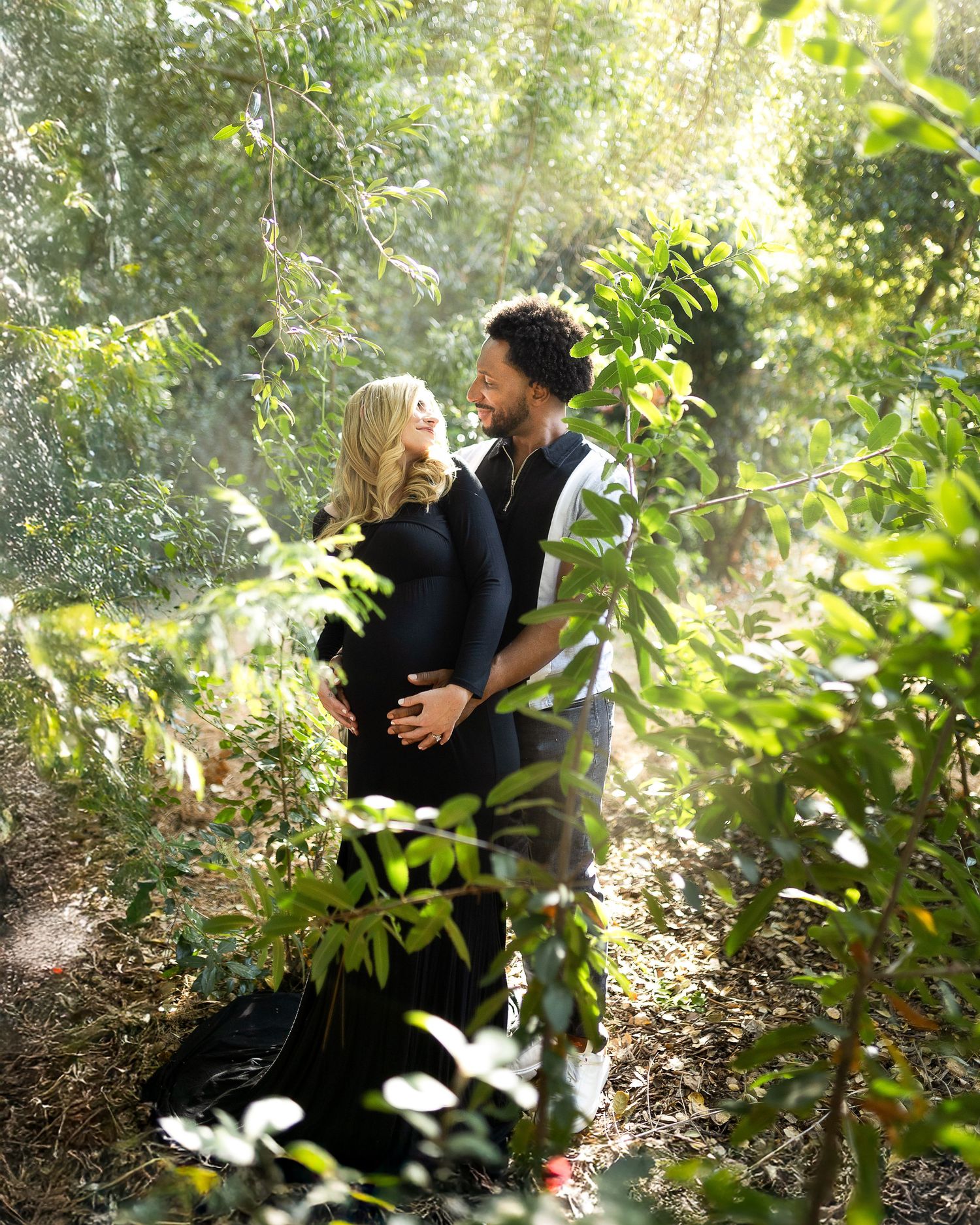 Intimate couple shares tender moments among trees and leaves in a sunlit forest setting.