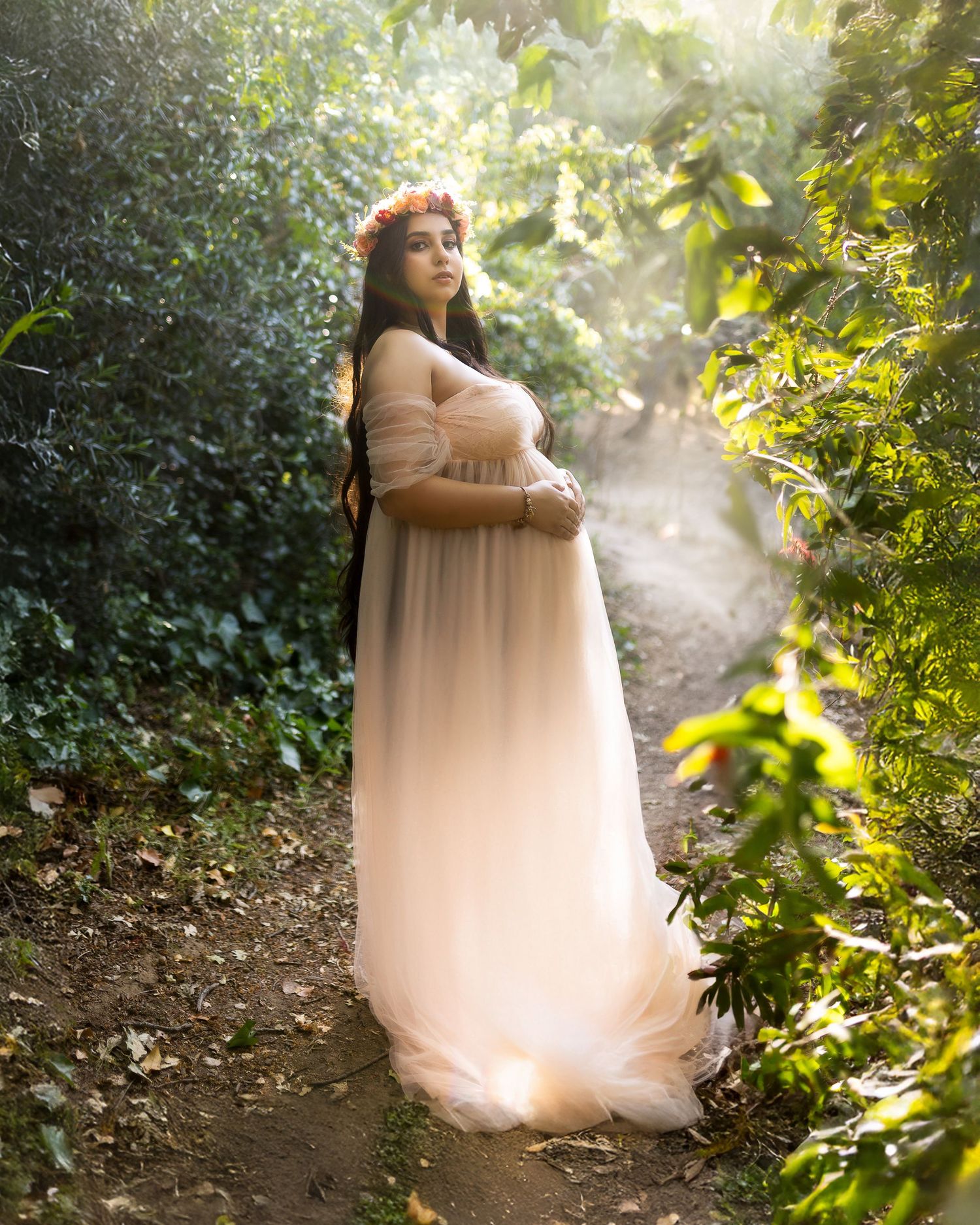 A pregnant woman in a flowing blush gown and flower crown poses ethereally on a sunlit forest path.