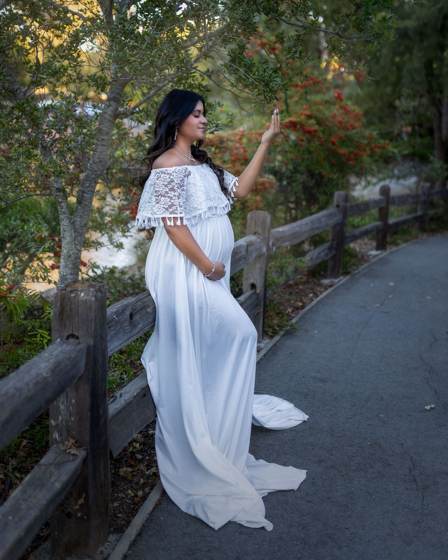 A figure in a flowing white dress poses gracefully along a rustic wooden railing in a scenic outdoor setting.